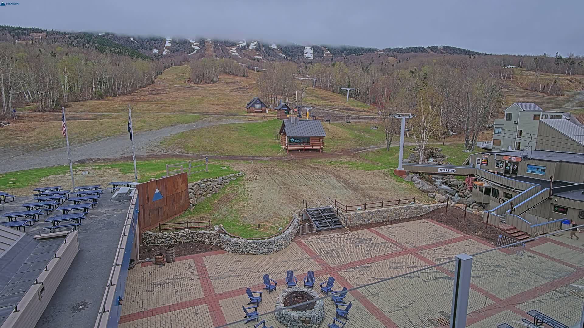 A paved outdoor area with picnic tables, a fire pit, and small buildings overlooks a ski resort with a few remaining patches of snow on the slopes under an overcast sky.
