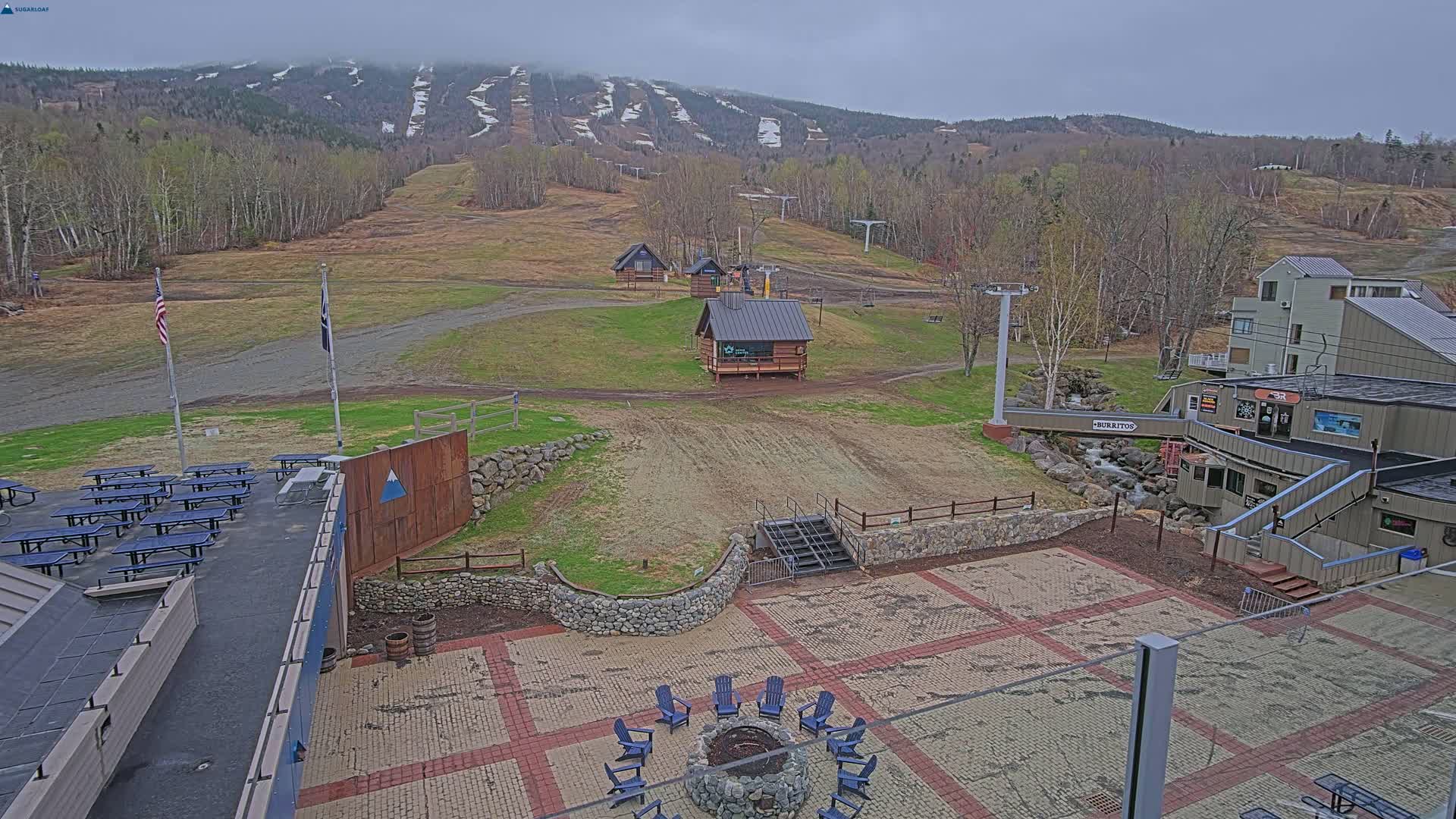 An outdoor patio area with picnic tables, a fire pit, and small buildings overlooks a ski slope with patchy snow, under an overcast sky.