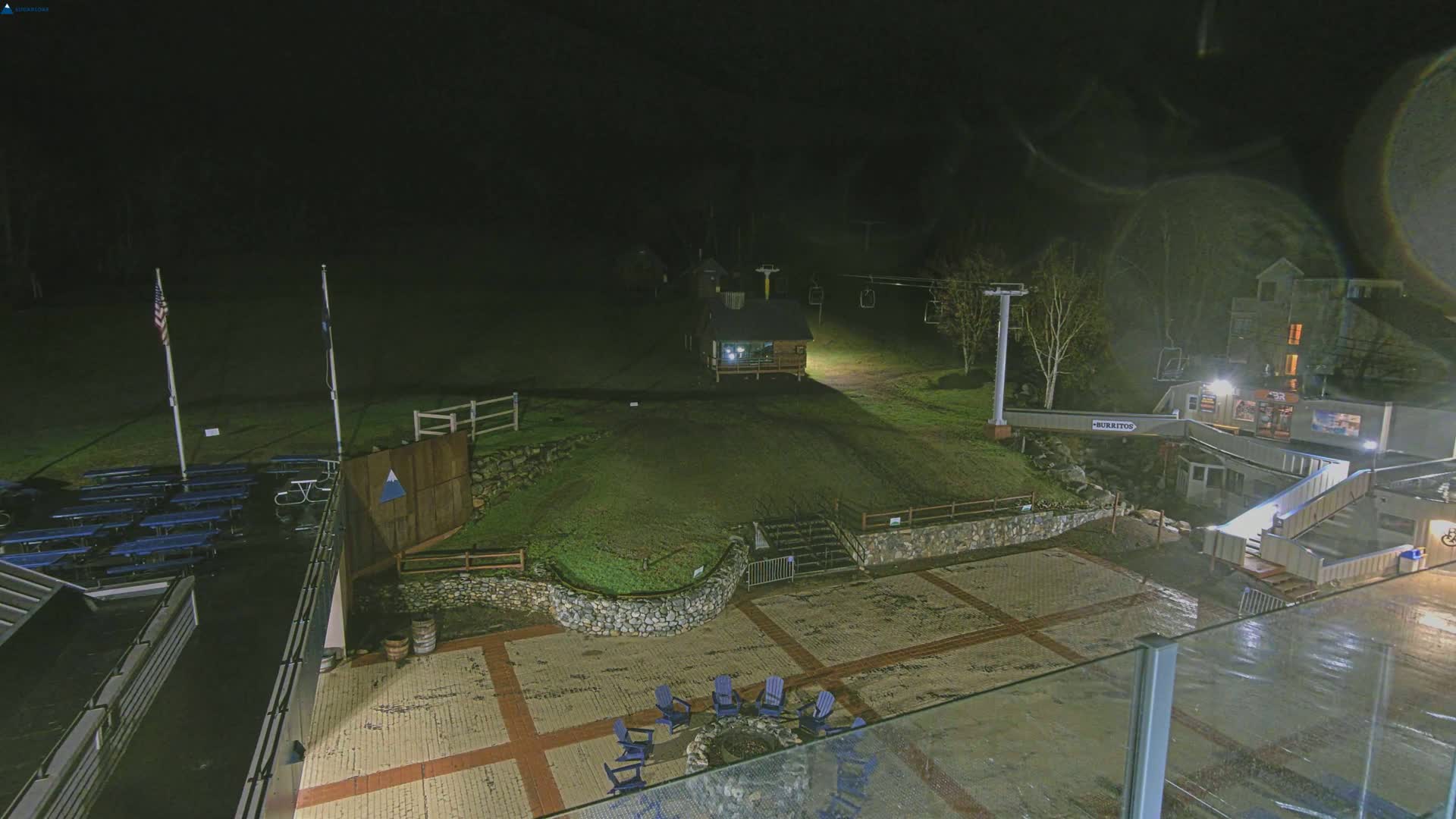 A nighttime view of a paved outdoor area with a fire pit and chairs, adjacent to a ski lift and buildings, under dark, wet conditions.