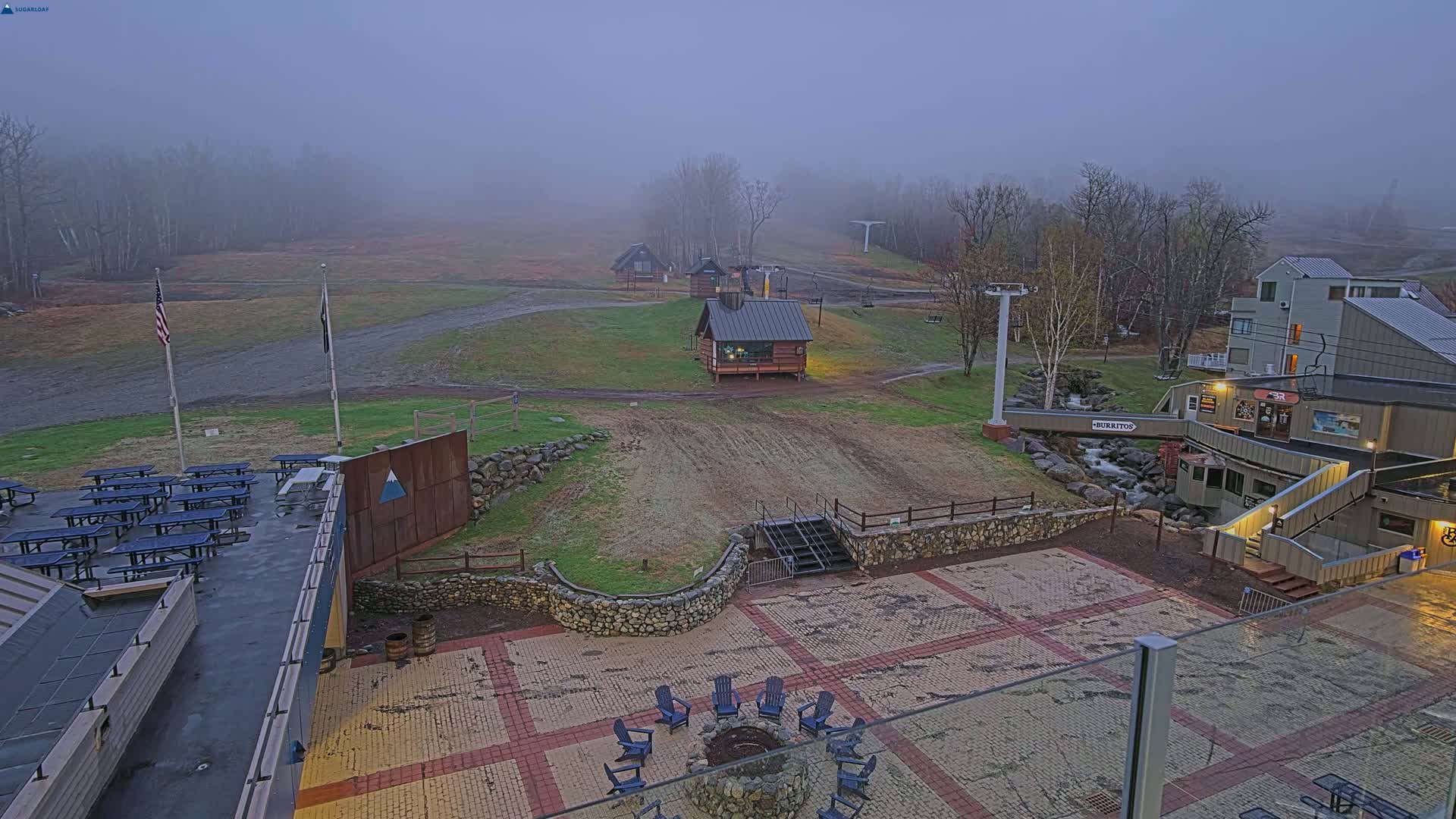 A foggy view of a ski resort base area shows a paved patio with a fire pit and Adirondack chairs, picnic tables, small buildings, and a ski lift in the background.