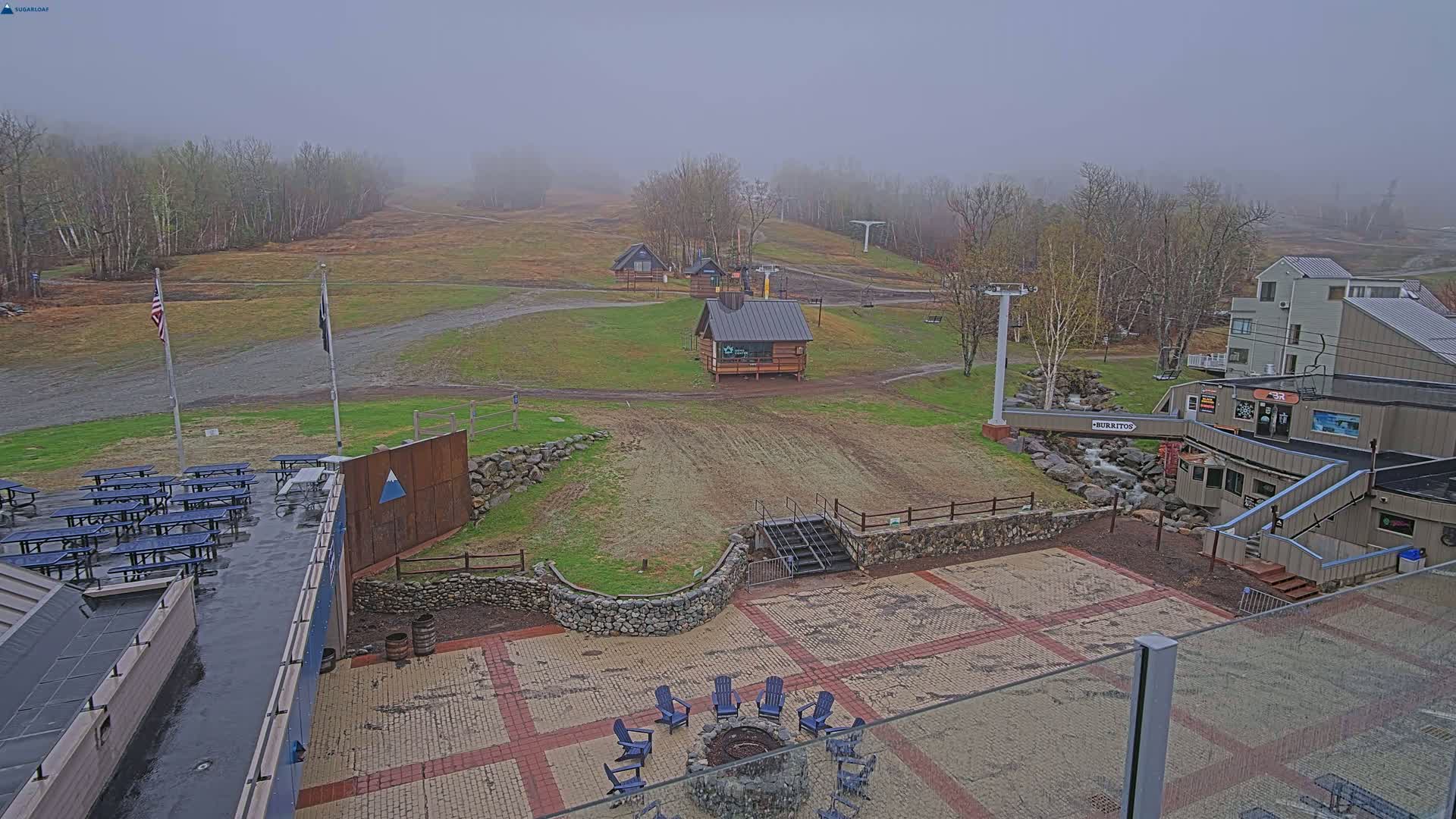 A misty outdoor scene shows a ski resort base area with a paved patio, picnic tables, a fire pit, several small buildings, a ski lift, and a grassy hillside.