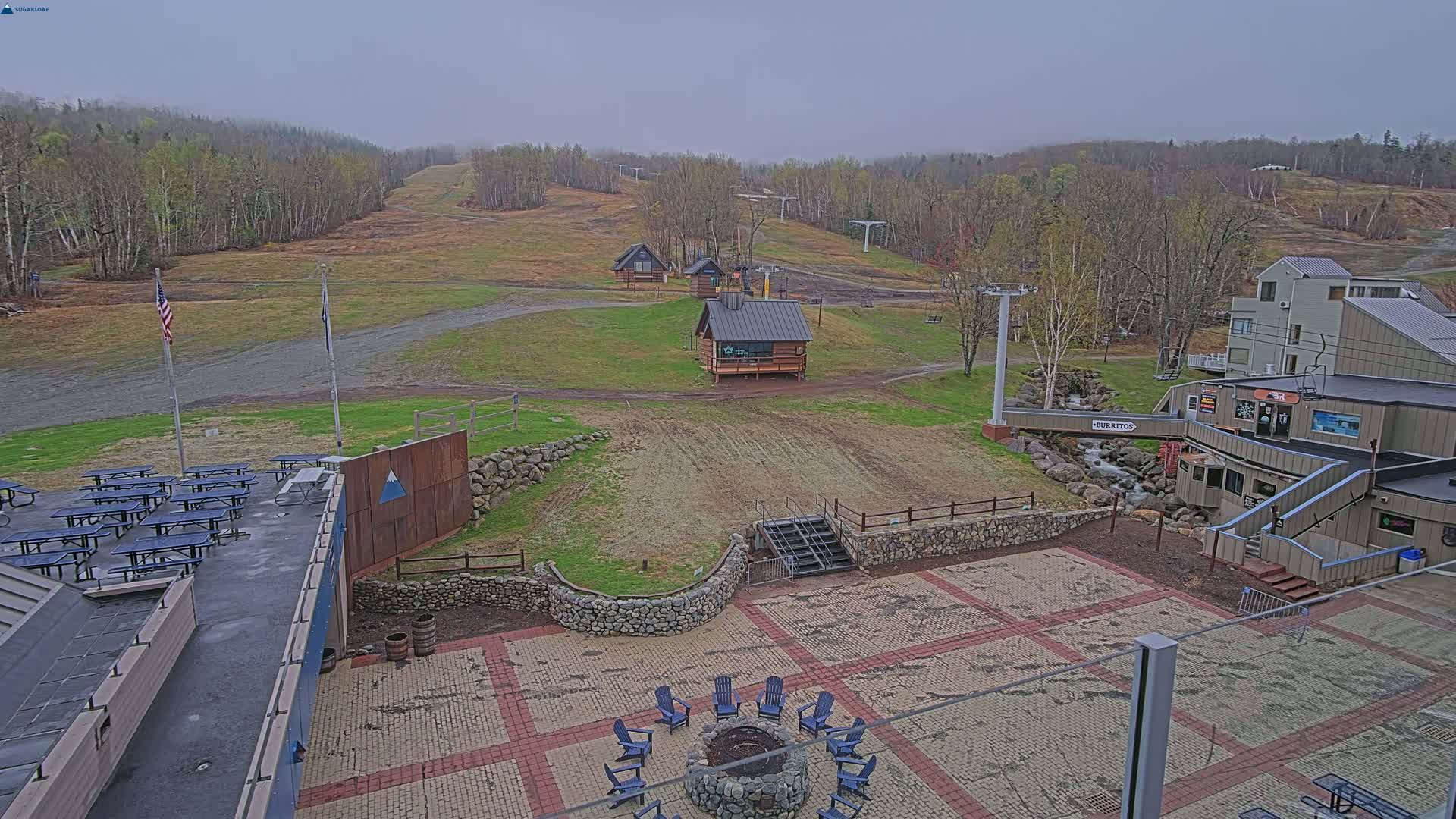 An overcast day reveals a ski resort's base area with picnic tables, a fire pit with chairs, and buildings, all situated amidst grassy slopes and ski lifts.
