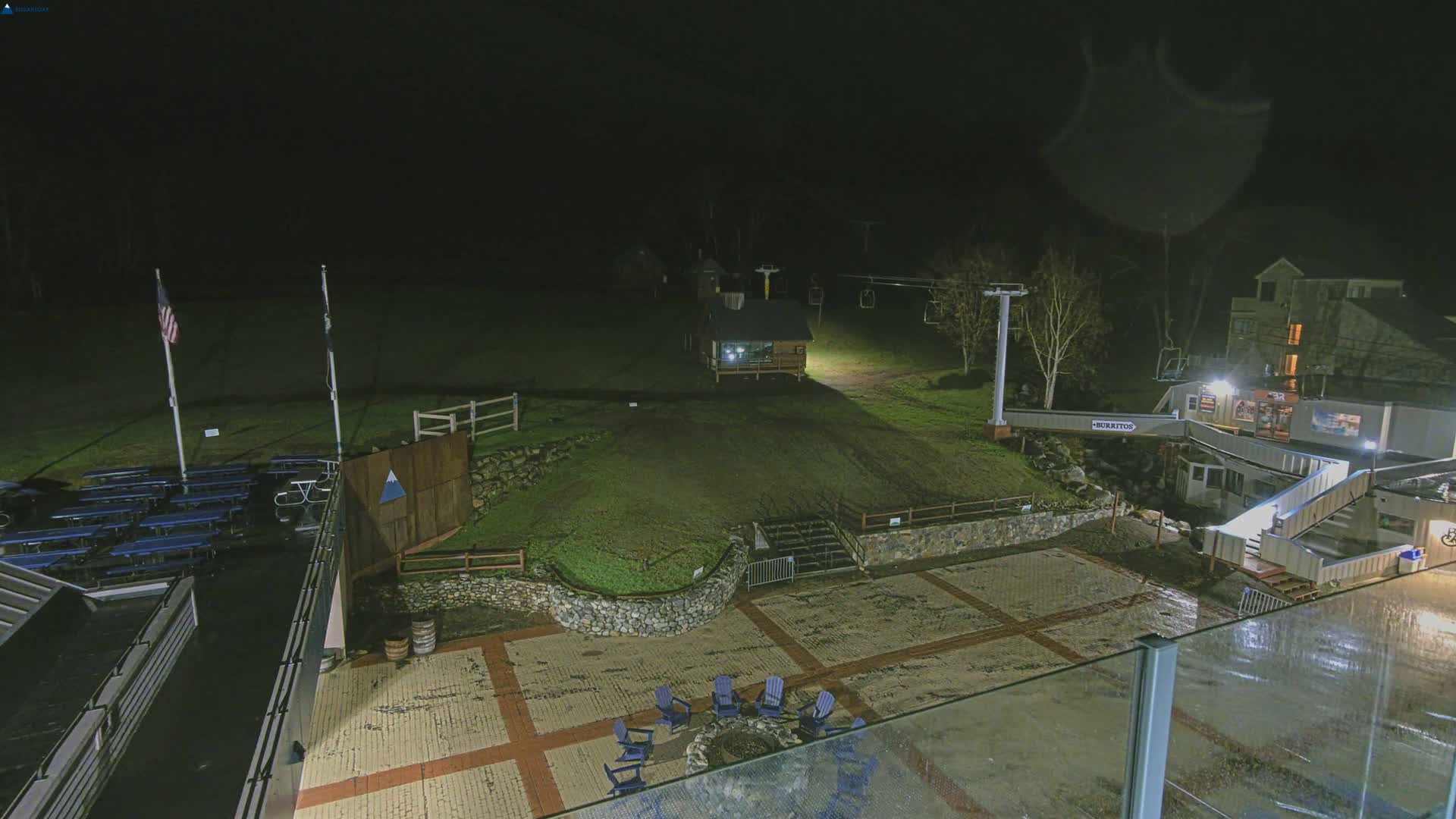 A nighttime view of an outdoor plaza area with a ski lift in the background, under wet conditions.