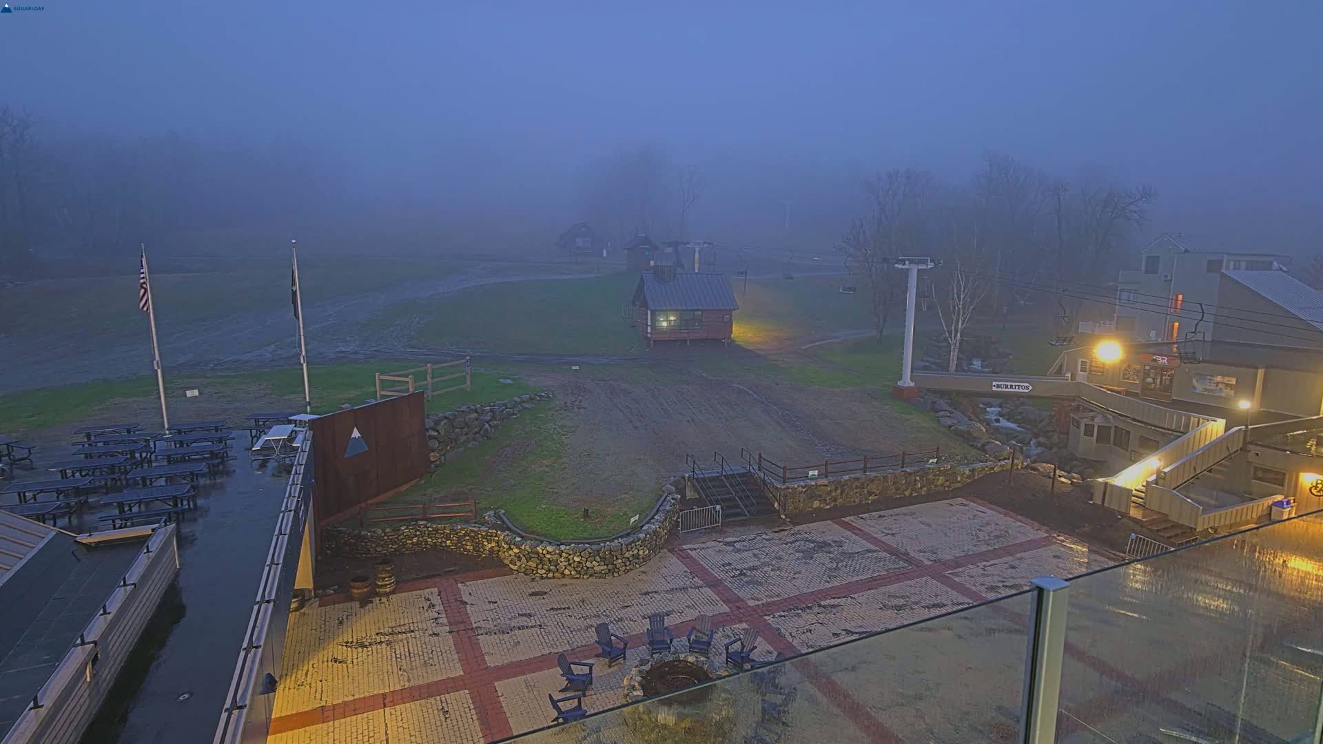 A foggy, dimly lit outdoor patio area with picnic tables, a fire pit, and buildings in the background.