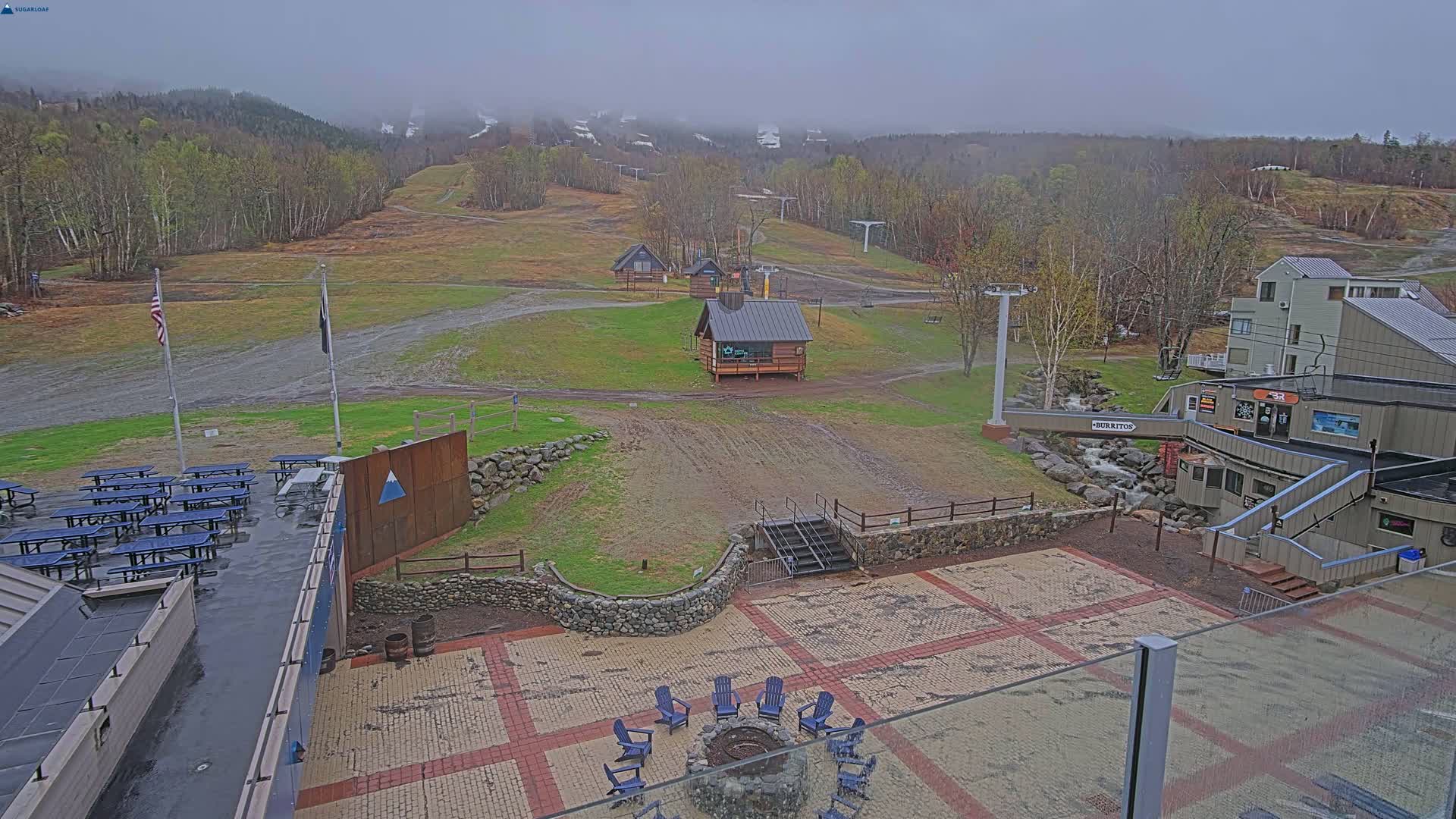 A misty, overcast day at a ski resort shows a paved patio area with picnic tables, a fire pit, and small buildings, overlooking grassy ski slopes and distant, fog-covered mountains.