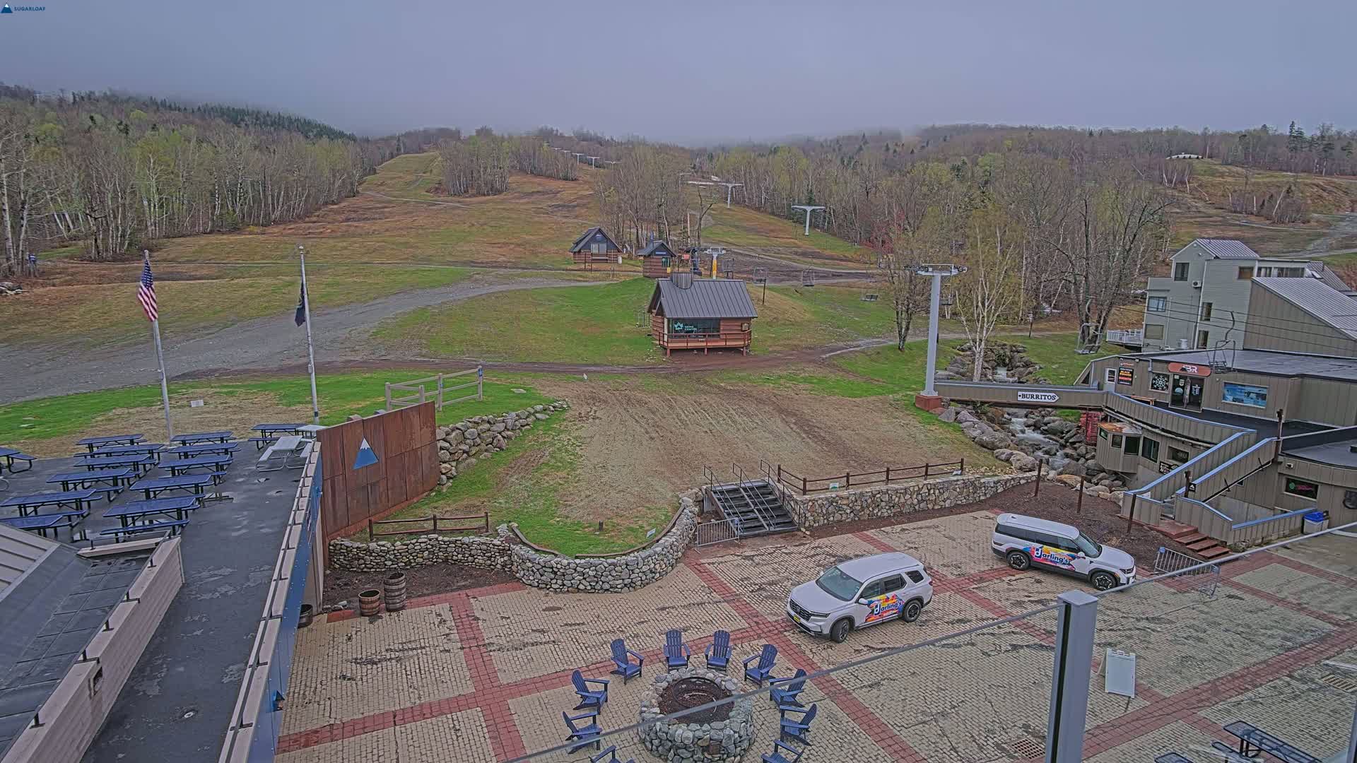 An overcast day at a ski resort shows a paved area with picnic tables, a fire pit, and two cars parked near a building, all overlooking grassy ski slopes and wooded hills in the distance.