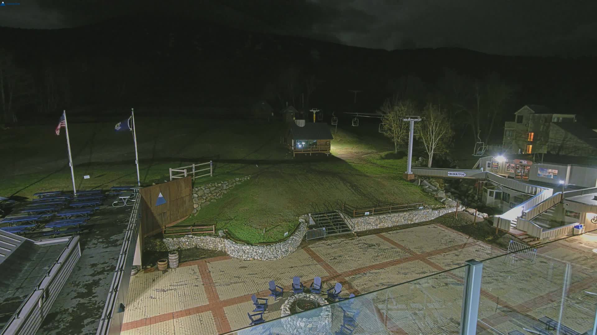 A nighttime view of a ski resort base area, showing a paved plaza with chairs around a fire pit, a small building, a ski lift, and American and state flags.