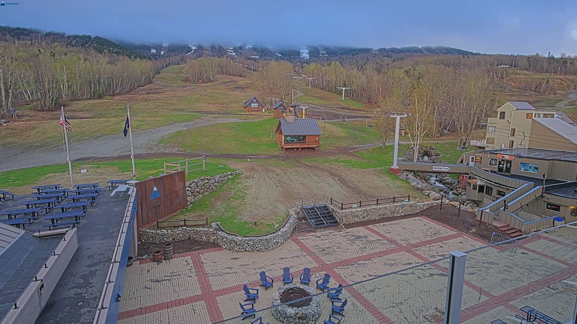 An outdoor patio area with picnic tables, a fire pit, and several small buildings overlooks a ski slope with trees and mountains in the background under an overcast sky.