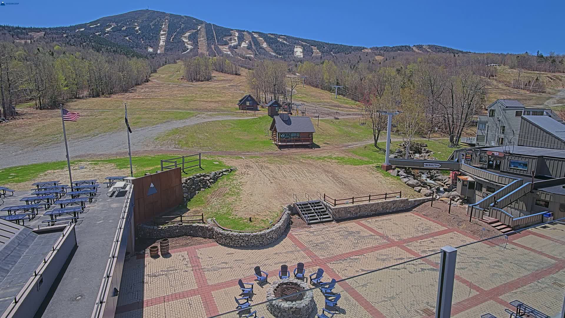 An outdoor patio area with a fire pit and picnic tables overlooks a ski resort with a mountain in the background under a clear, sunny sky.