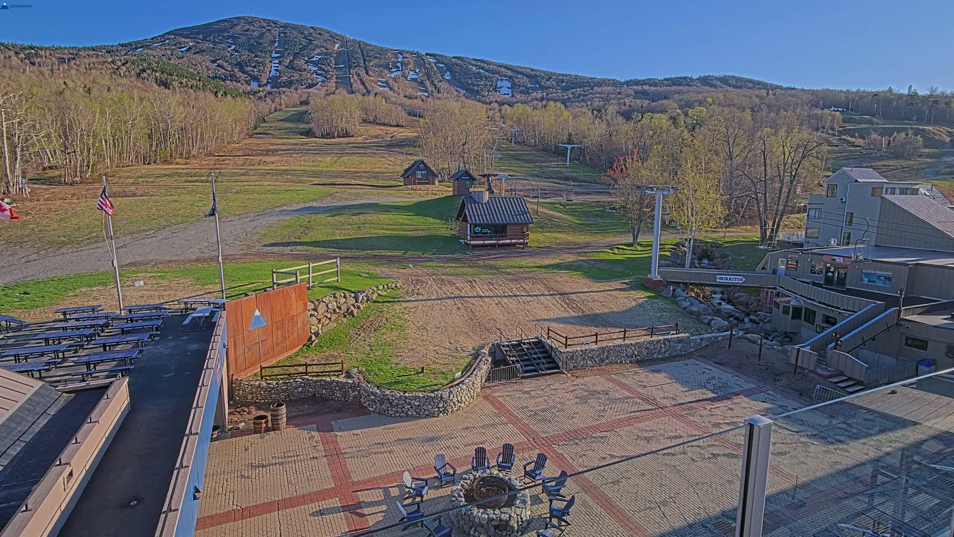 An outdoor patio area with a fire pit and picnic tables overlooks a ski slope and mountain under a clear blue sky.