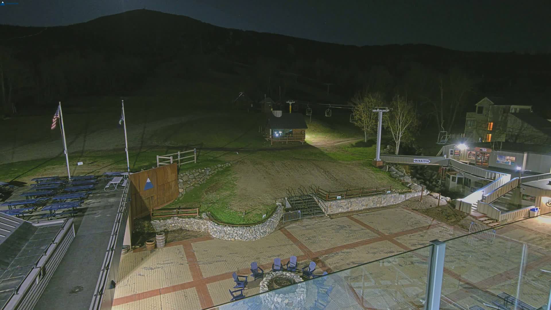 A nighttime view of a ski resort patio area with a fire pit, picnic tables, a chairlift, and a building in the background under a clear, moonlit sky.