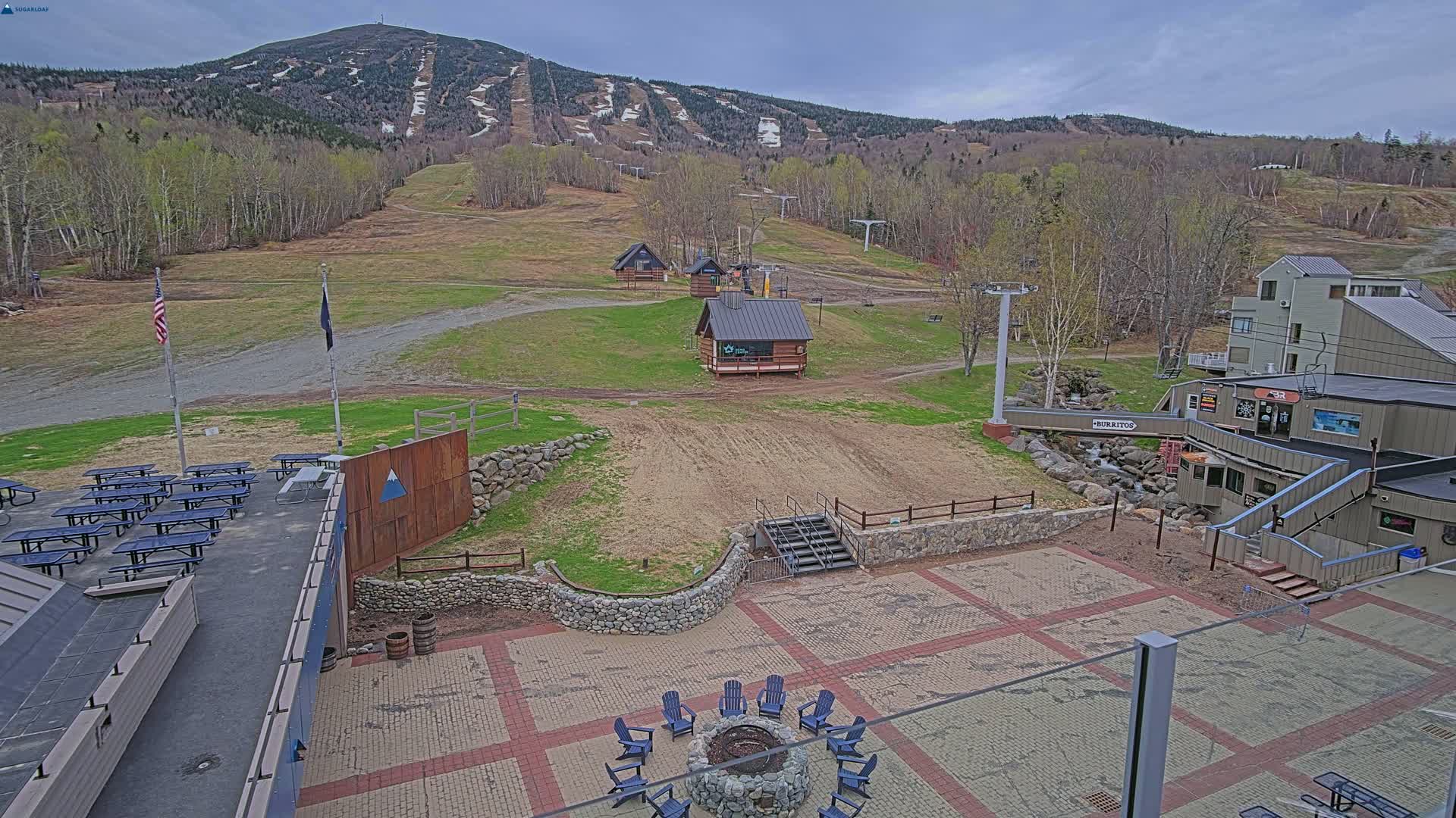 An overcast day reveals a ski resort with several buildings, a paved patio area with a fire pit and picnic tables, and a mountain in the background with visible ski trails.