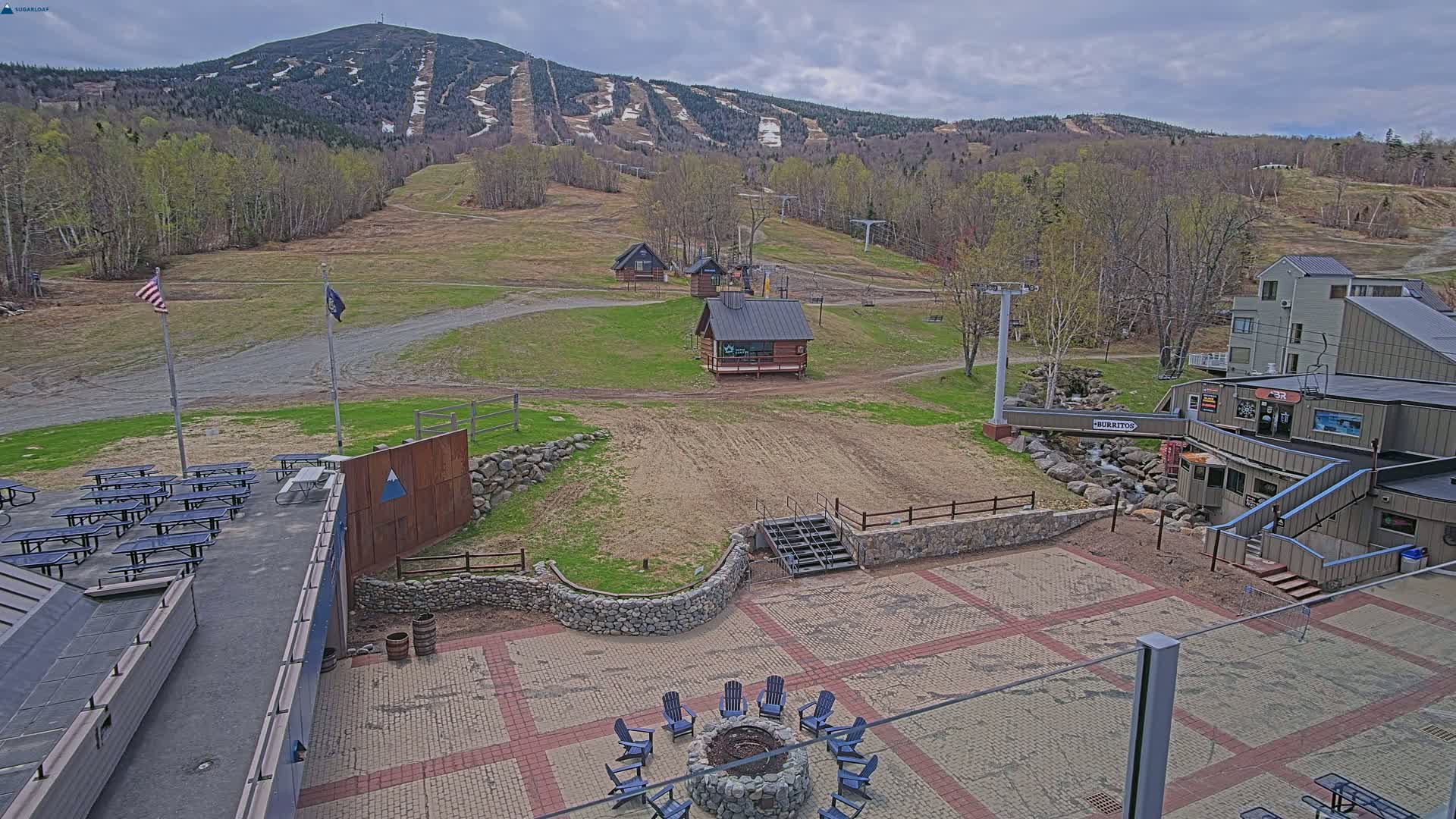 A paved outdoor area with picnic tables, a fire pit with chairs, and small buildings overlooks a ski resort with mostly bare slopes and budding trees under a partly cloudy sky.
