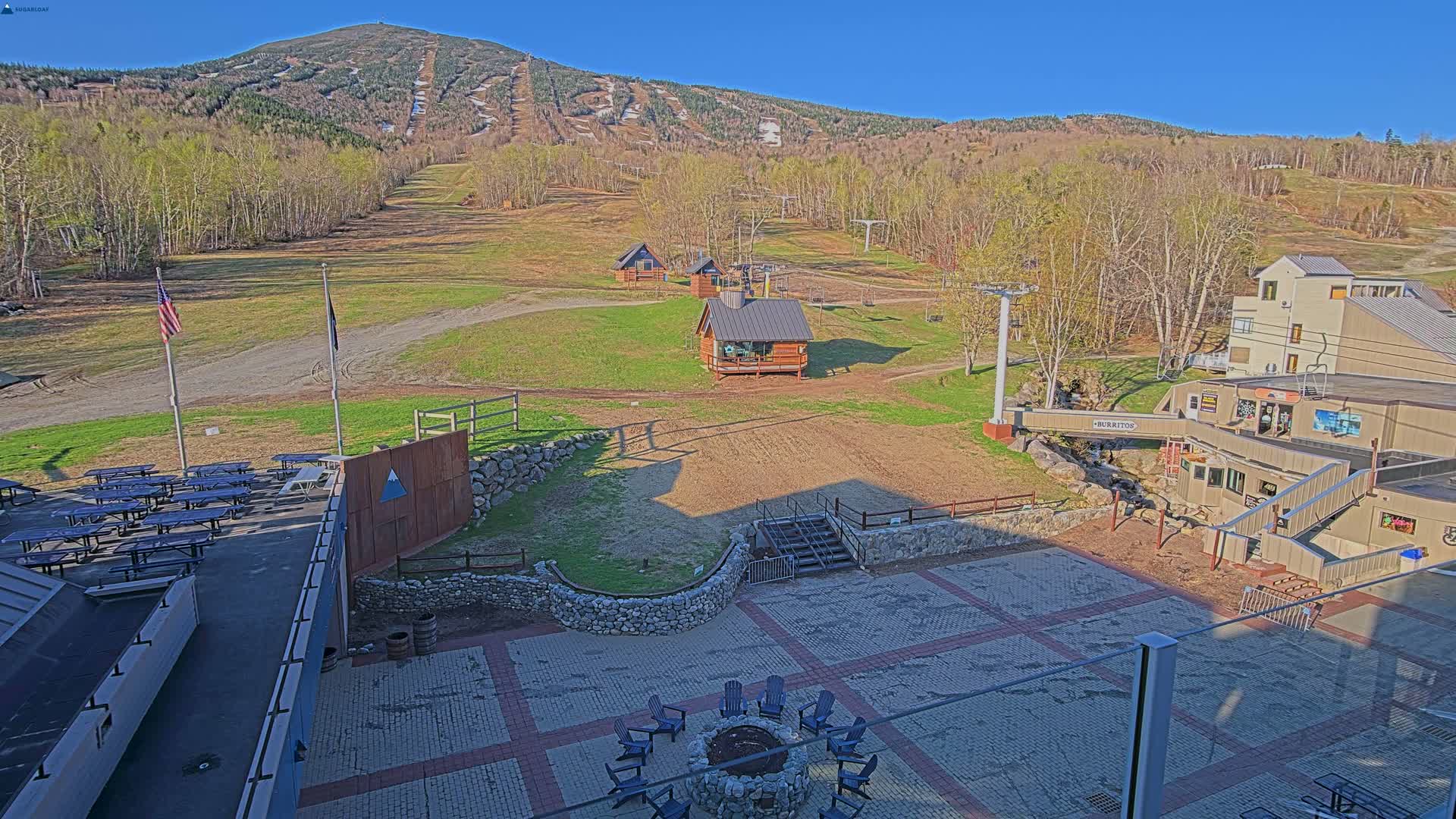 A sunny outdoor scene shows a ski resort with grassy slopes, small buildings, a paved patio area with picnic tables and a fire pit, and a mountain in the background.