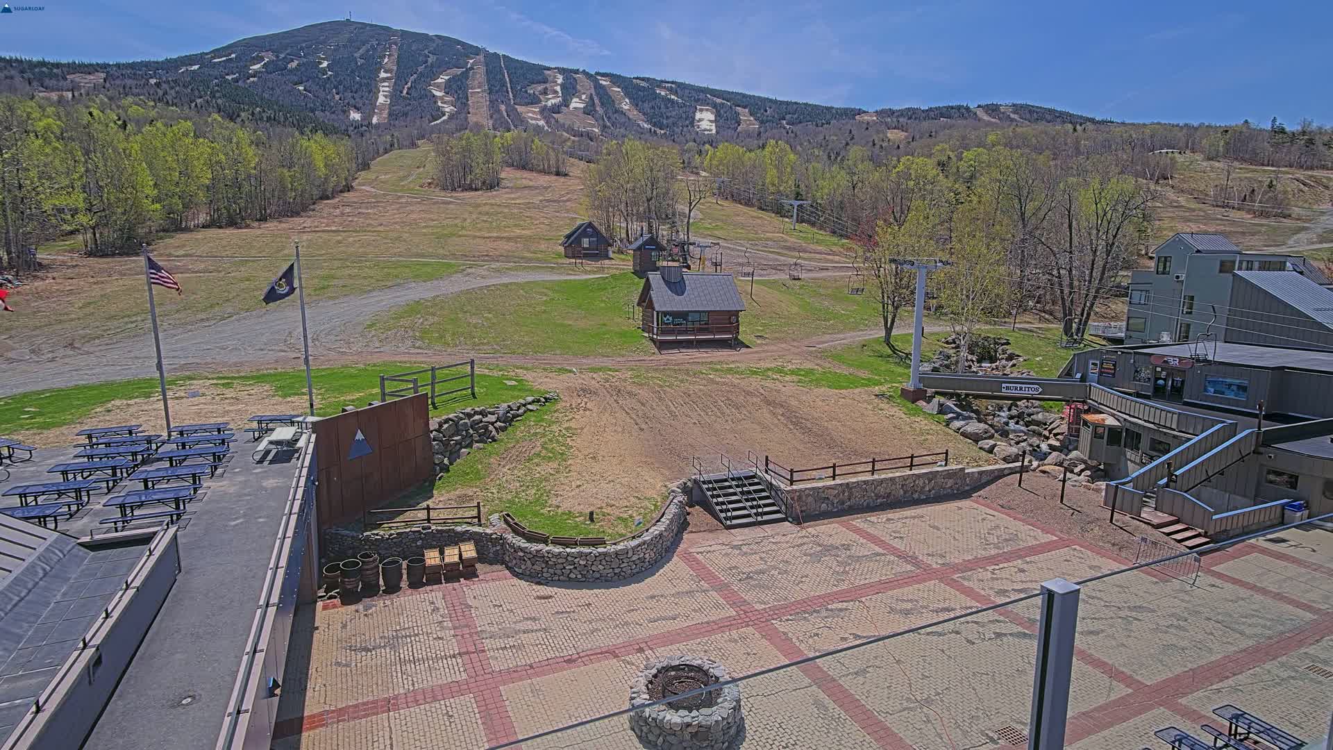 A paved outdoor area with picnic tables and a stone fire pit overlooks a ski resort with grassy slopes, several small buildings, and a mountain in the background on a sunny day.