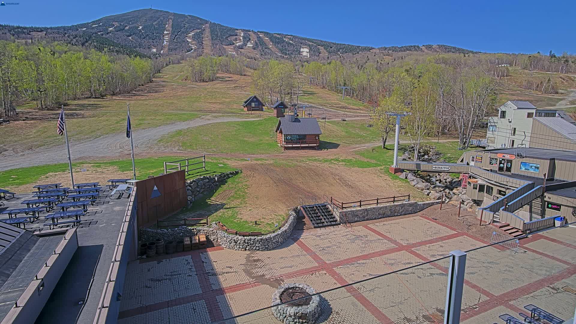 An outdoor patio area with picnic tables, small buildings, and a ski lift overlooks a grassy ski slope and mountain with patches of snow under a clear blue sky.