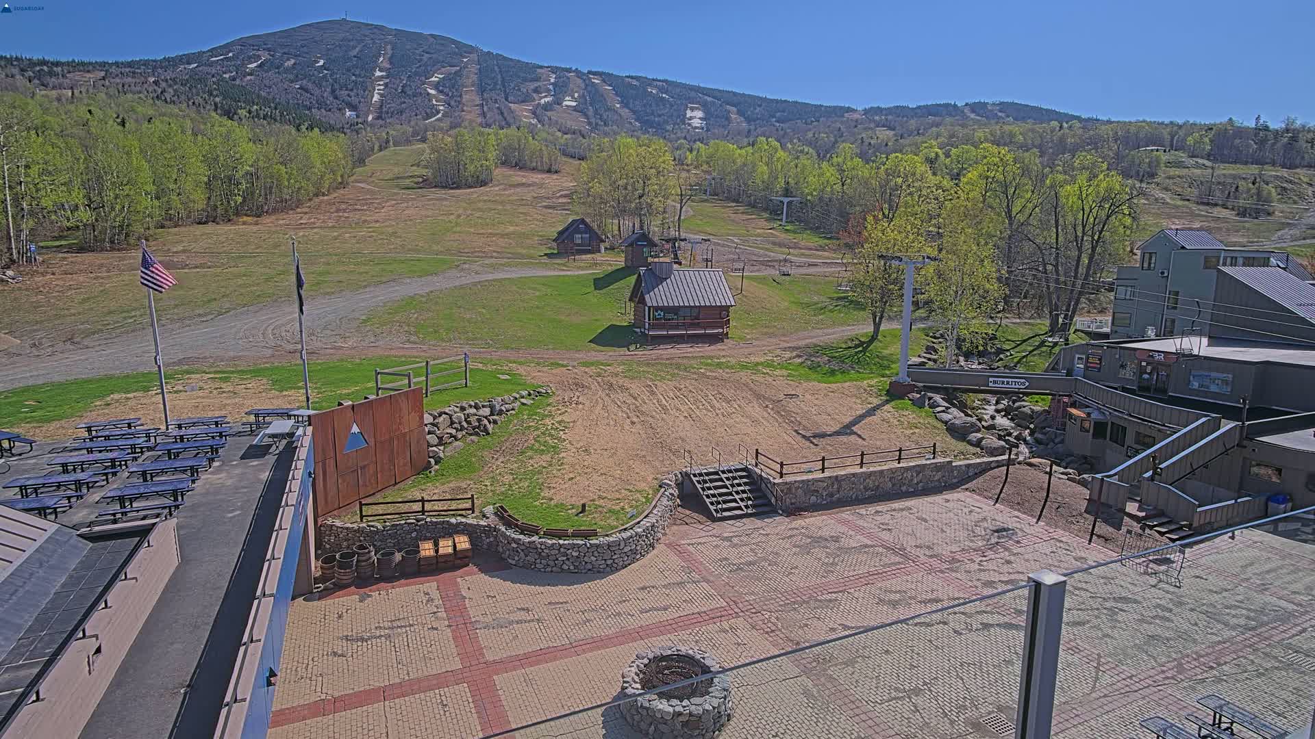 A paved patio area with picnic tables and small buildings sits below a ski slope on a sunny day, with a mountain in the background.