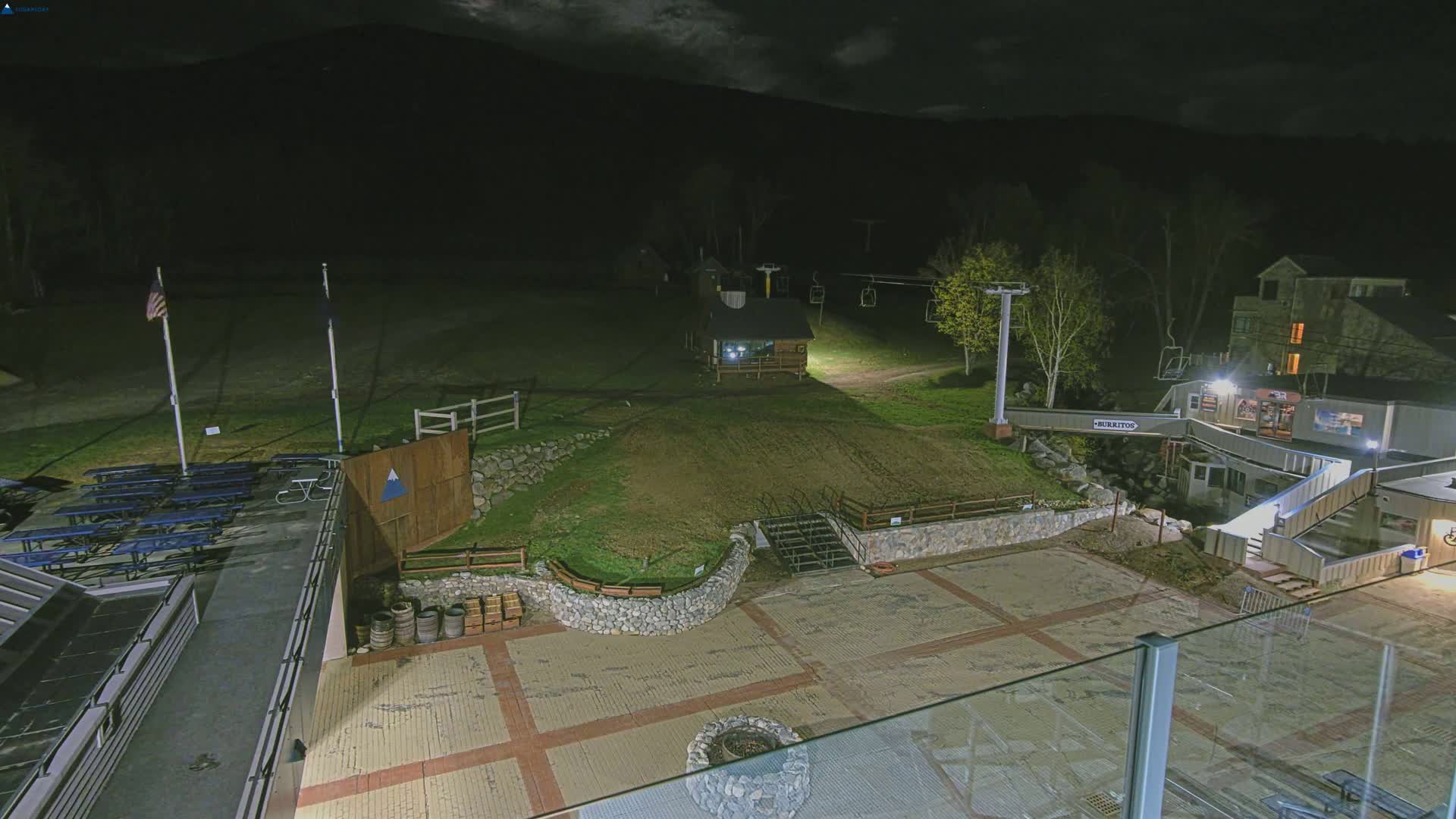 A nighttime view of a ski resort base area showing a paved plaza with picnic tables, a small building, a ski lift, and a stone retaining wall, all under a dark sky.