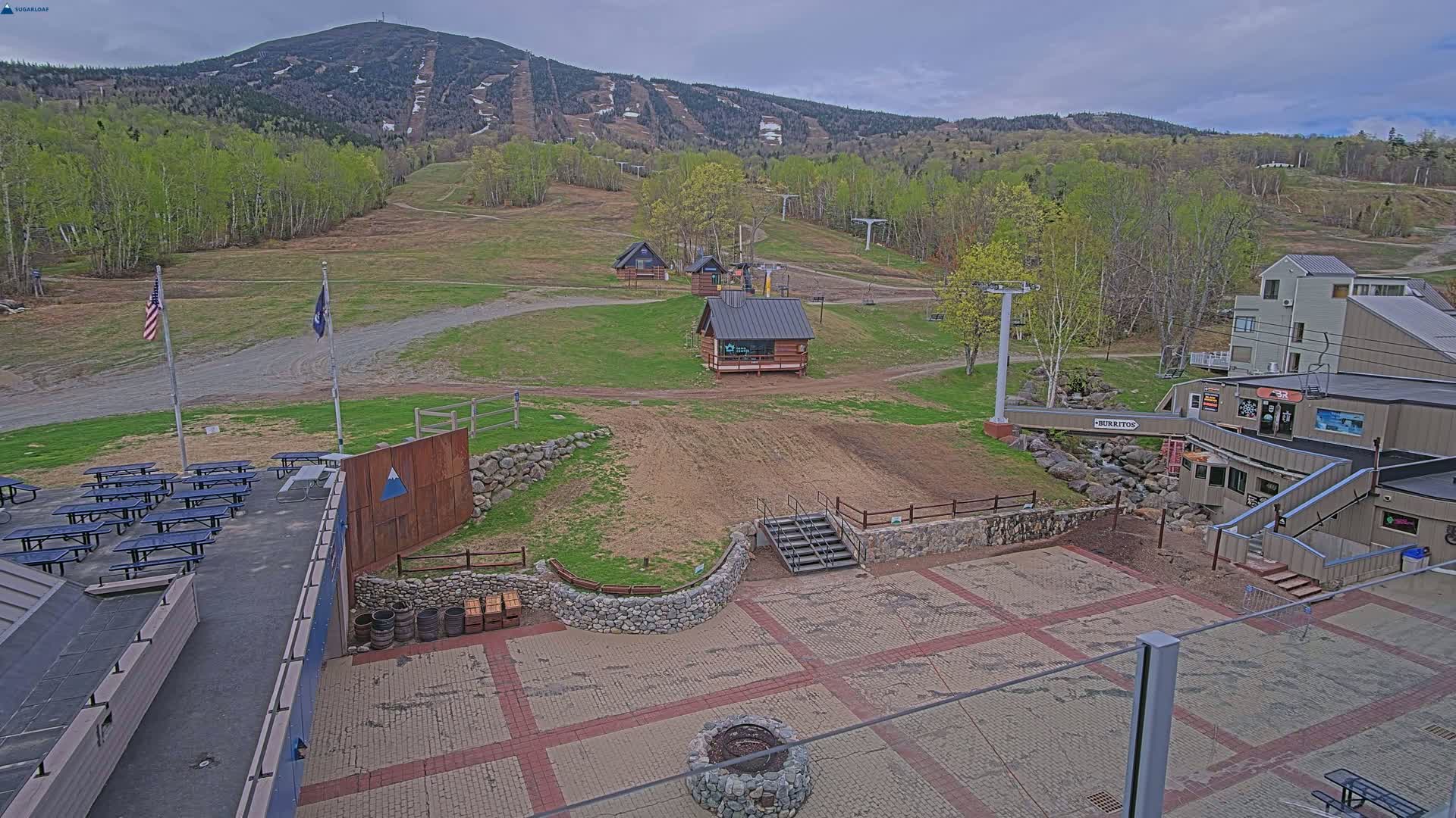 A paved area with picnic tables, small buildings, and a ski lift overlooks a grassy hillside with a mountain in the background under an overcast sky.