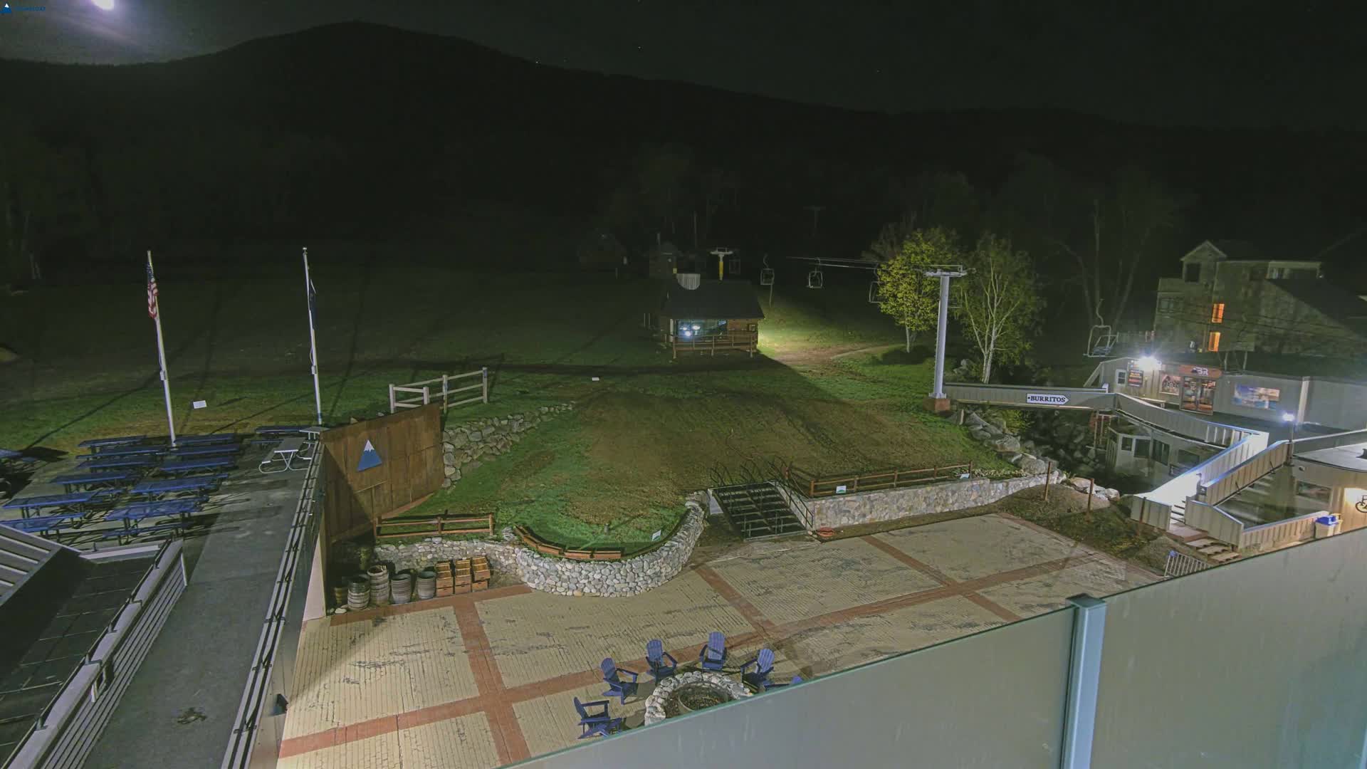 A nighttime view of a ski resort base area with a paved plaza, fire pit, picnic tables, a small building, ski lift, and a building in the background under a clear, moonlit sky.