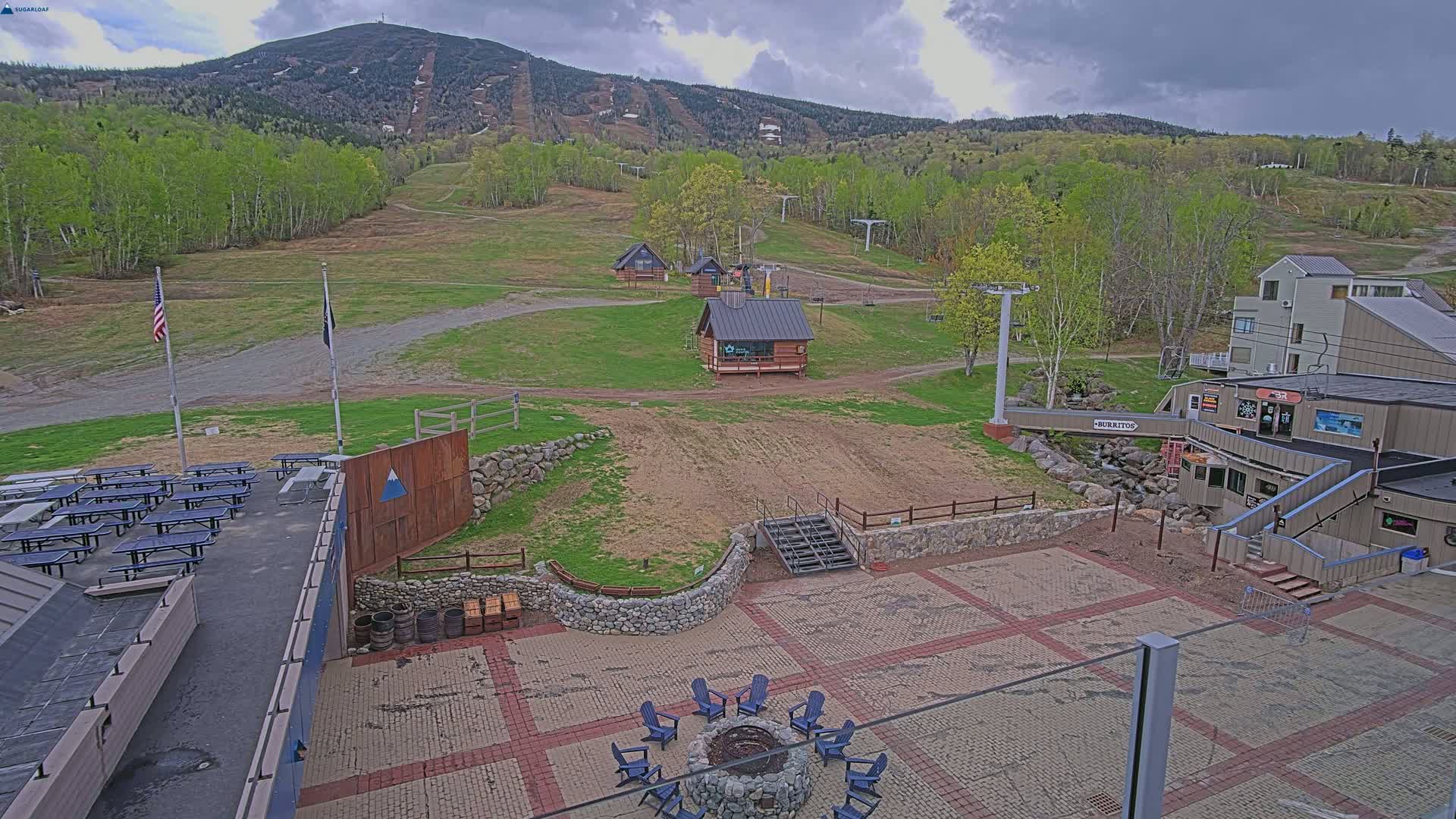 An outdoor paved patio area with a fire pit and seating, picnic tables, small buildings, ski lift infrastructure, and a mountain in the background under cloudy skies.