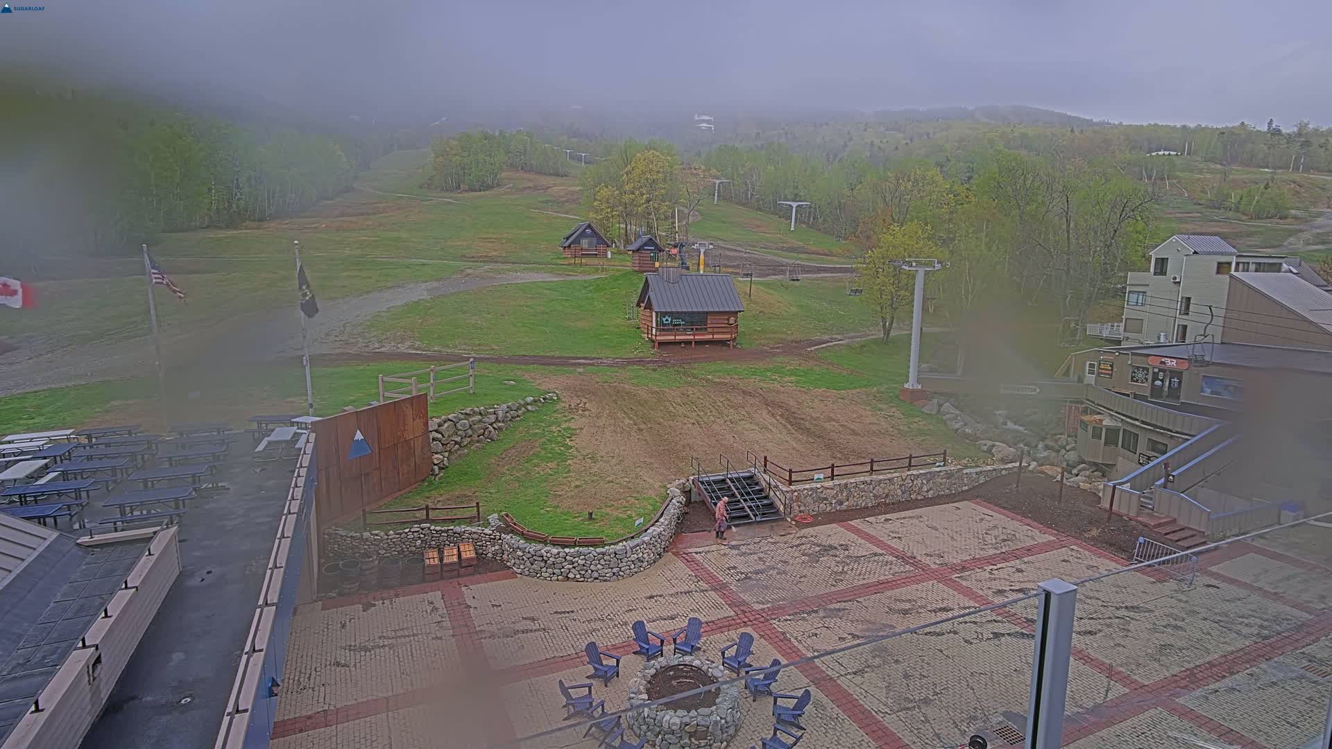 A foggy outdoor scene shows a ski resort patio with a fire pit and picnic tables, overlooking grassy ski slopes with small buildings and ski lifts.