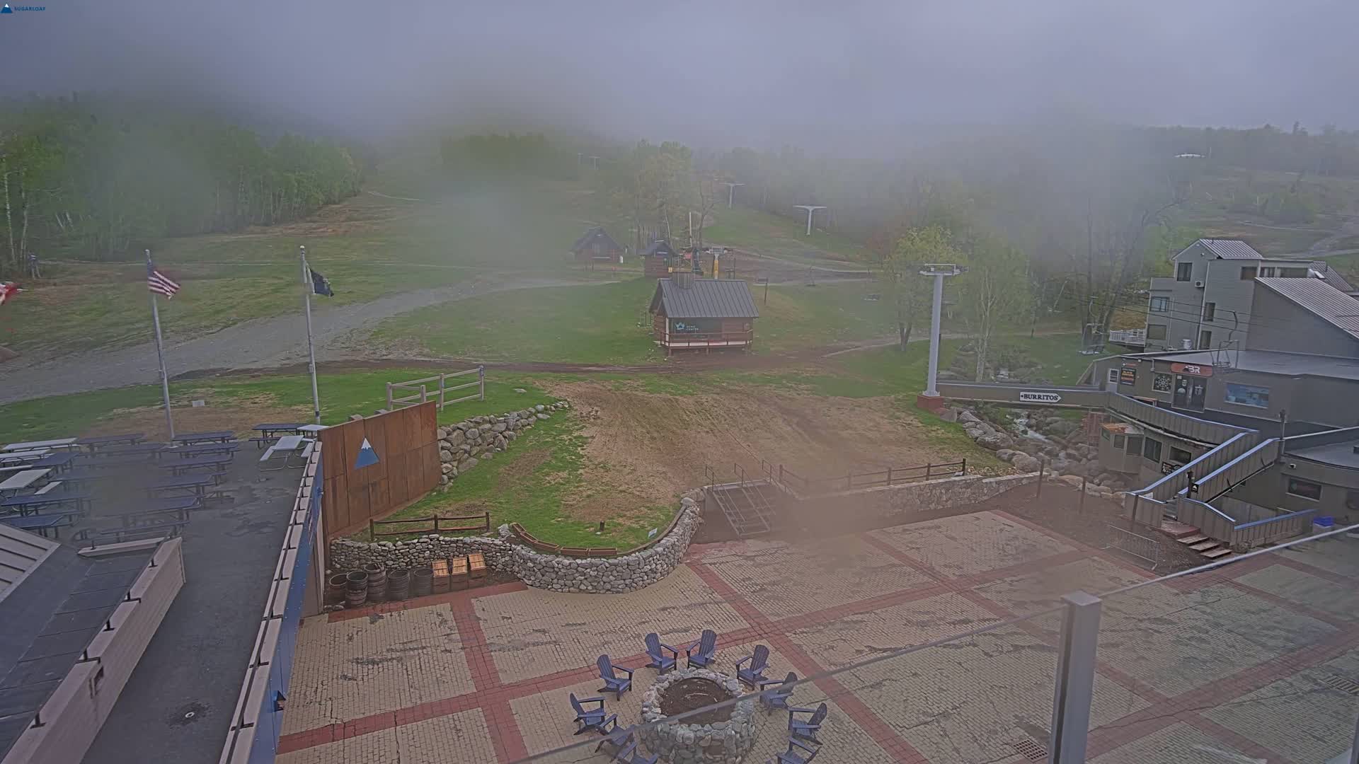 A foggy outdoor scene shows a paved area with a fire pit and chairs, surrounded by buildings, grassy areas, and ski lift infrastructure.