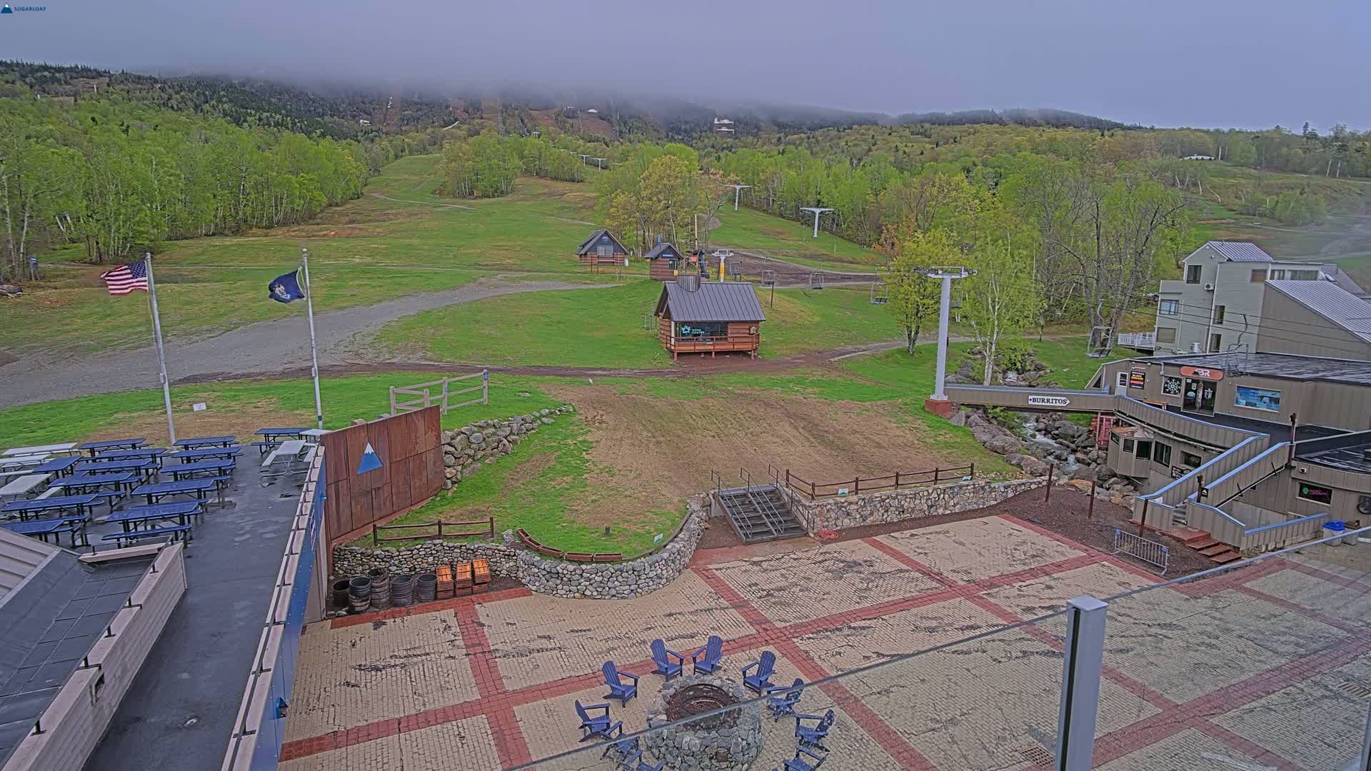 An overcast day reveals a ski resort's base area with grassy slopes, buildings, picnic tables, and a fire pit.