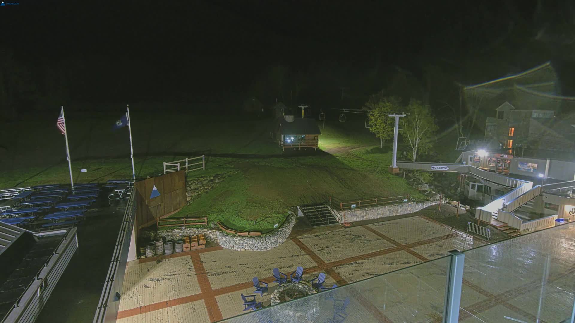 A nighttime view of an outdoor patio area with a stone wall, picnic tables, a small building, a chairlift, and flags, under dark, possibly rainy conditions.