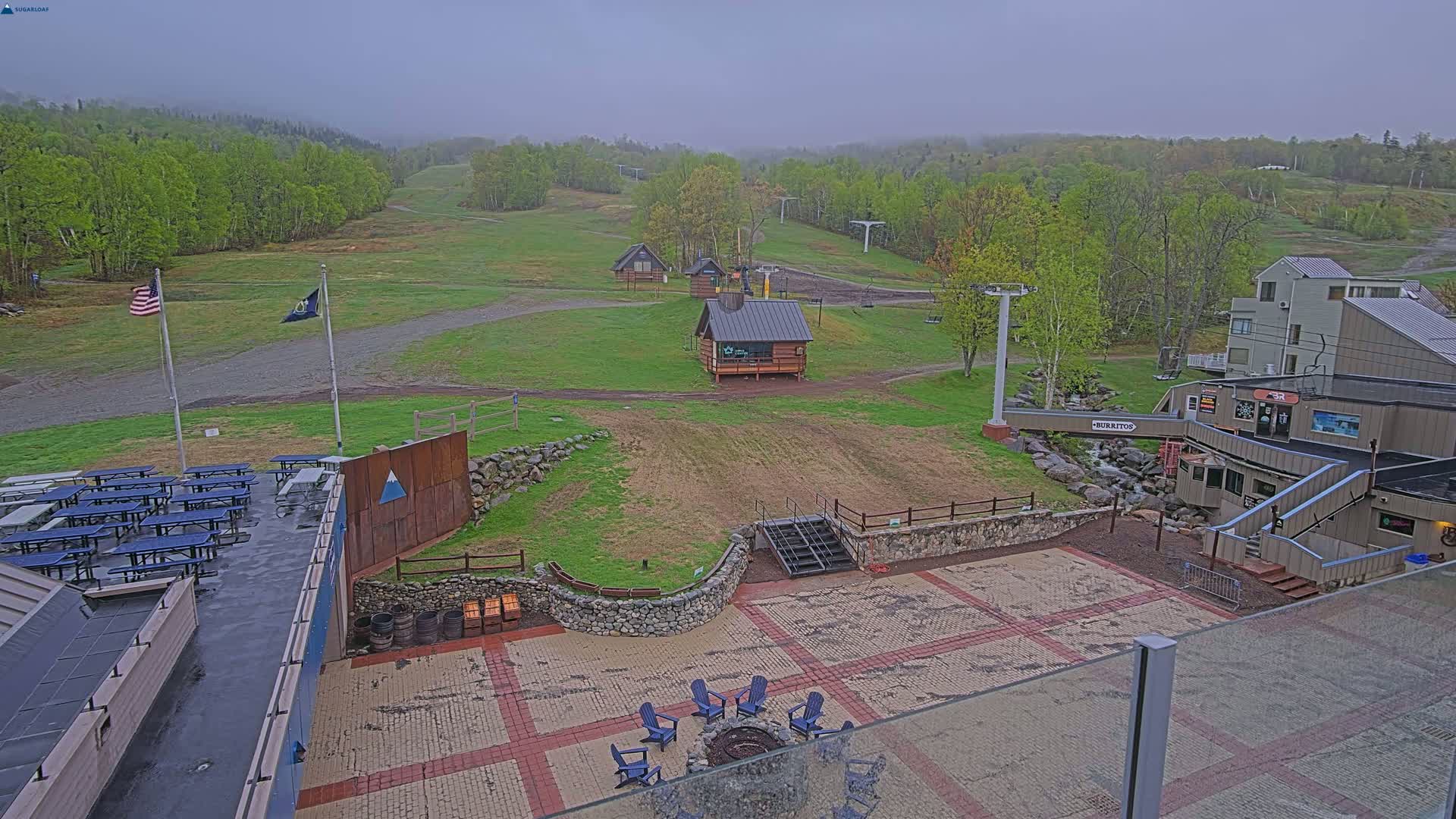 An outdoor patio area with a fire pit and seating overlooks a grassy ski slope with several small buildings under an overcast sky.
