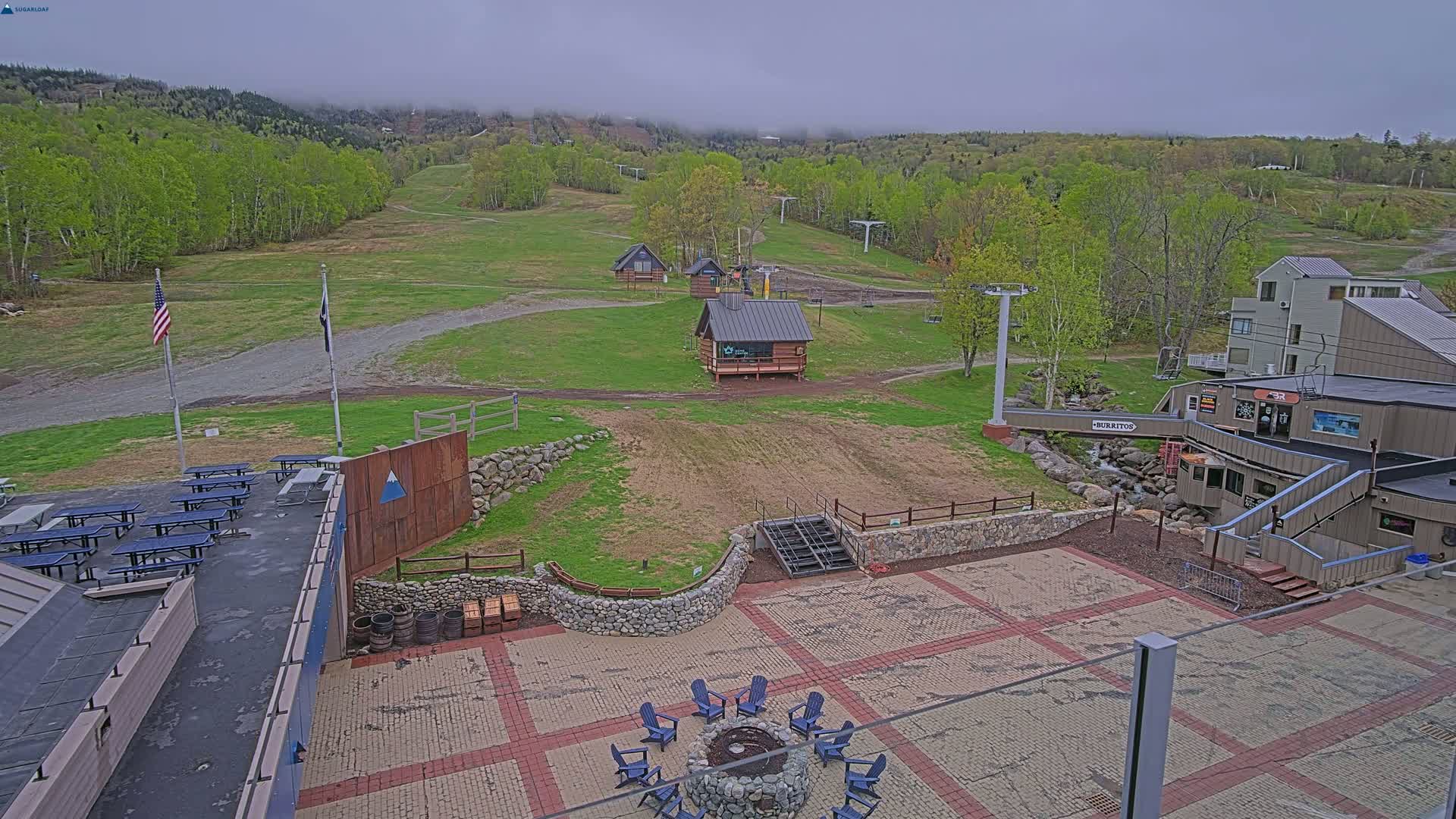 A ski resort base area with multiple buildings, picnic tables, a fire pit, and a paved patio, under an overcast sky.
