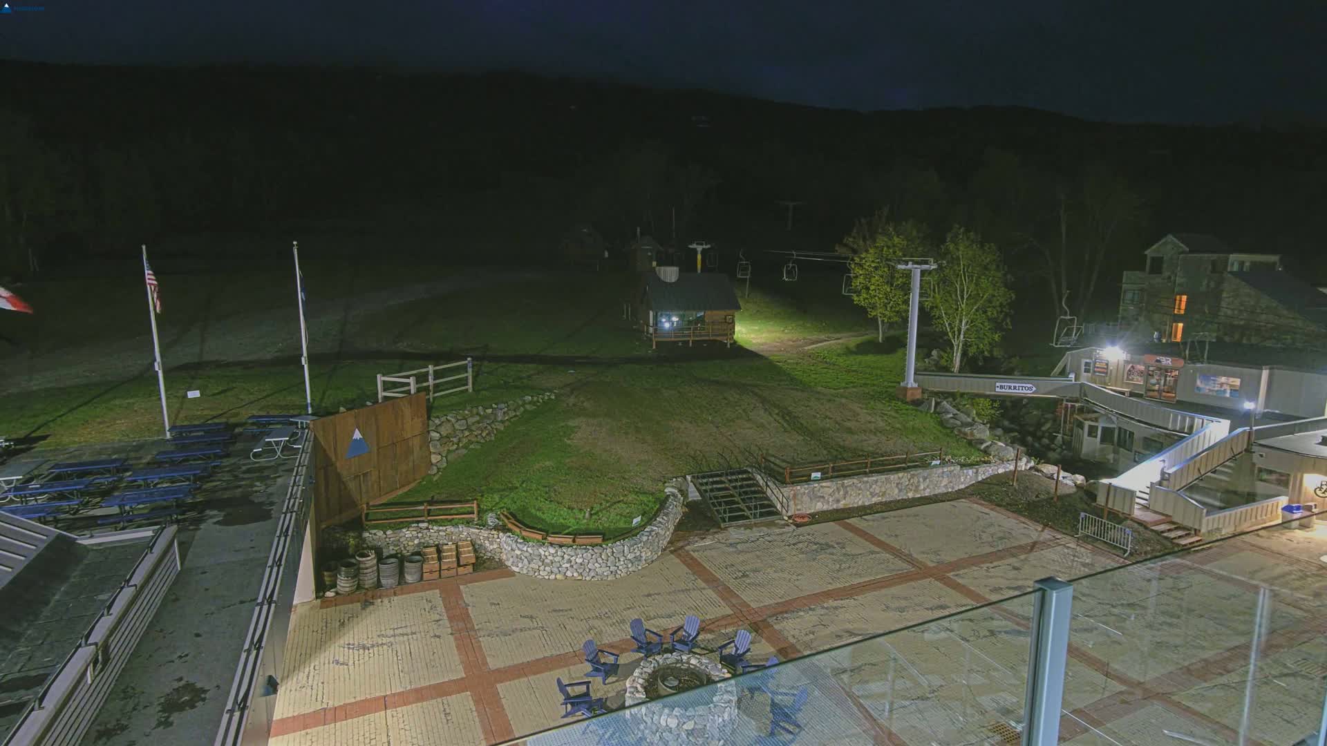 A nighttime view of an outdoor area with picnic tables, a fire pit, a small building, and a ski lift, illuminated against a dark, wooded hillside.