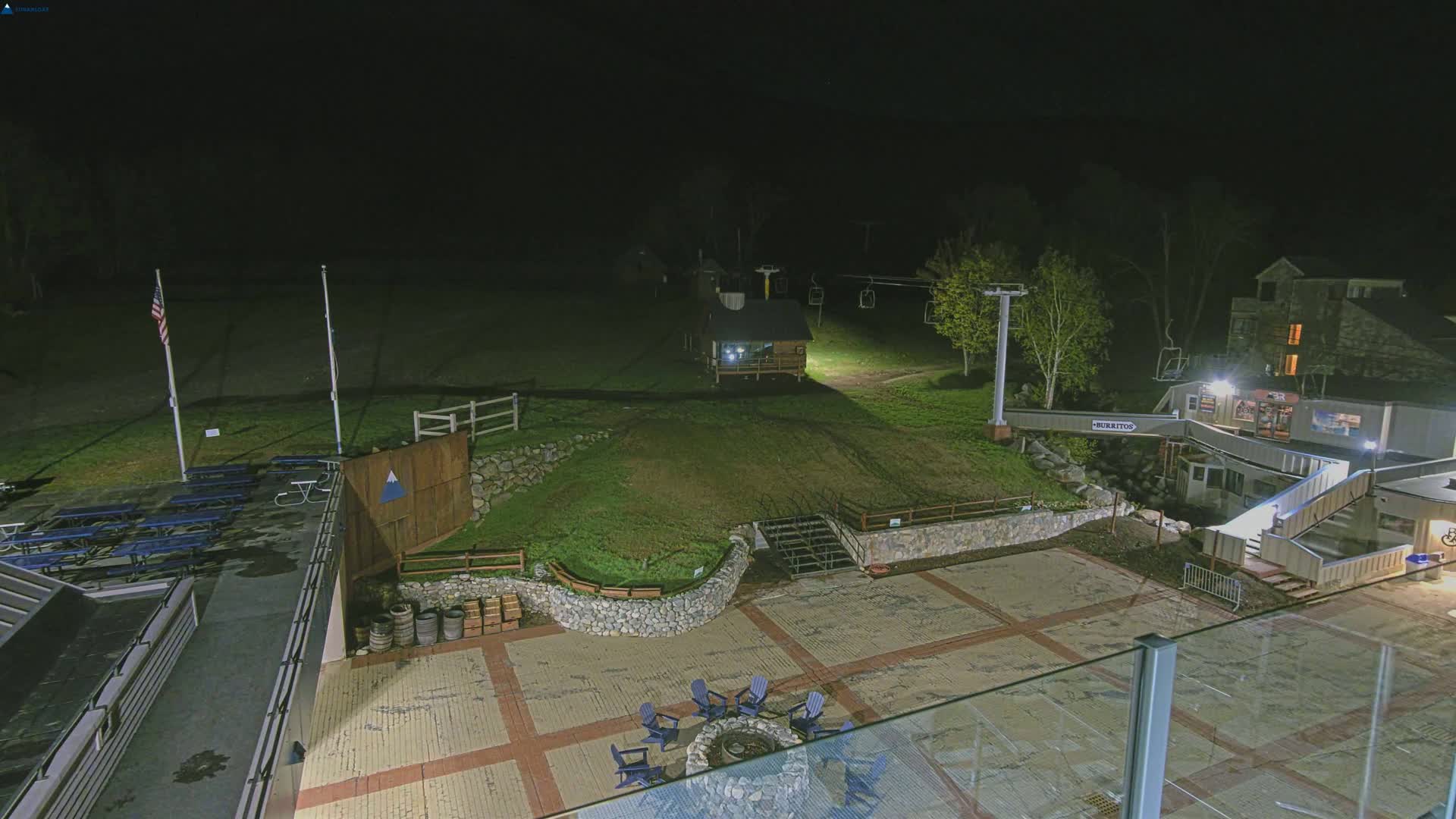 A nighttime view of an outdoor plaza area at a ski resort, featuring a fire pit, picnic tables, a small building, ski lifts, and a stone retaining wall.