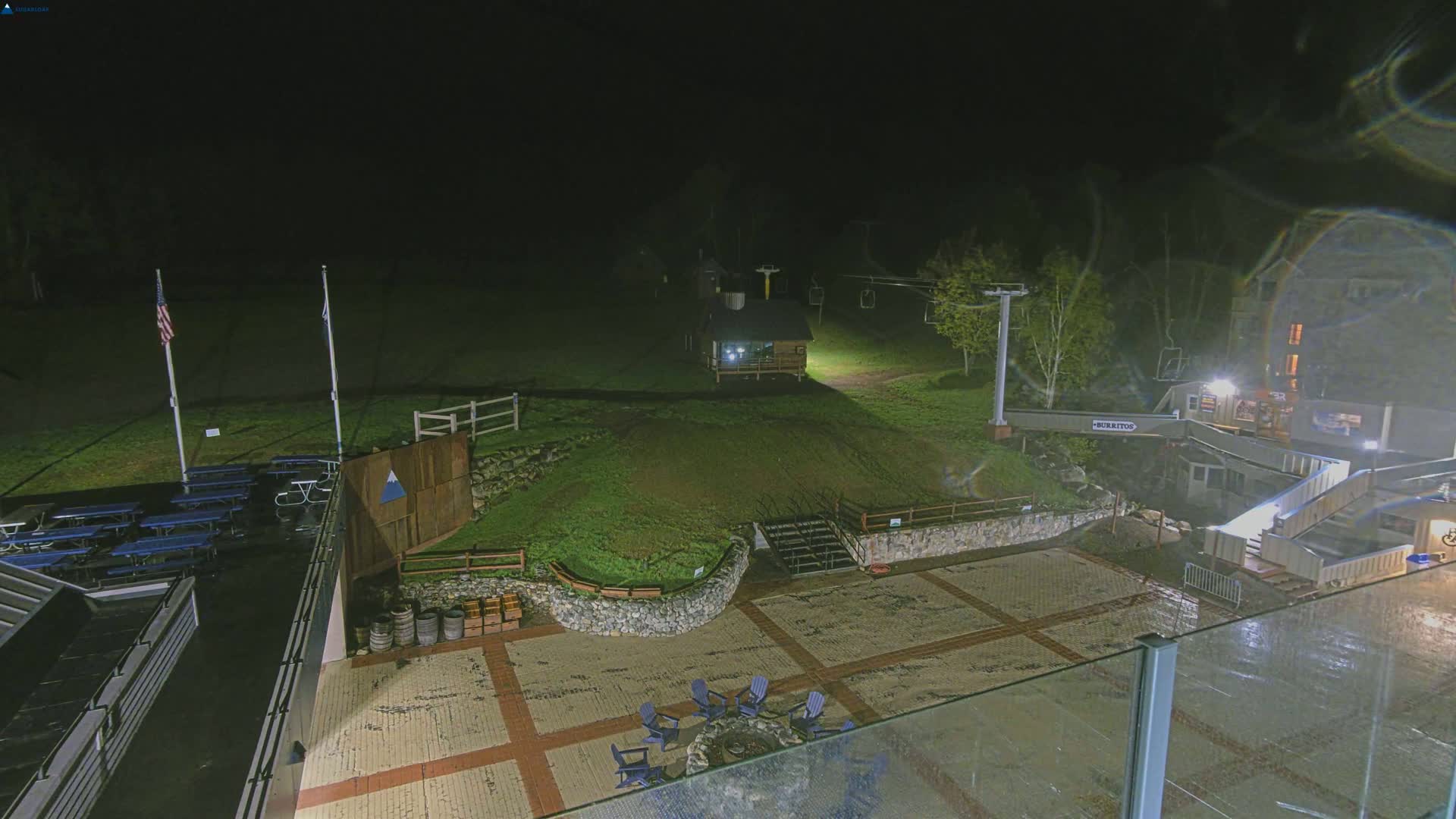 A nighttime view of an outdoor area with a paved plaza, picnic tables, a small building, a ski lift, and a fire pit, under dark conditions.