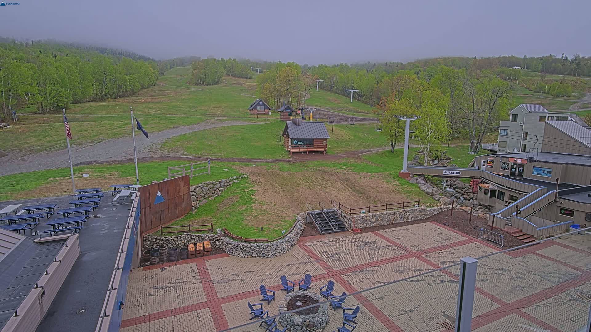 A paved patio area with picnic tables and a fire pit is situated below a ski lift and several small buildings, overlooking a grassy ski slope under an overcast sky.
