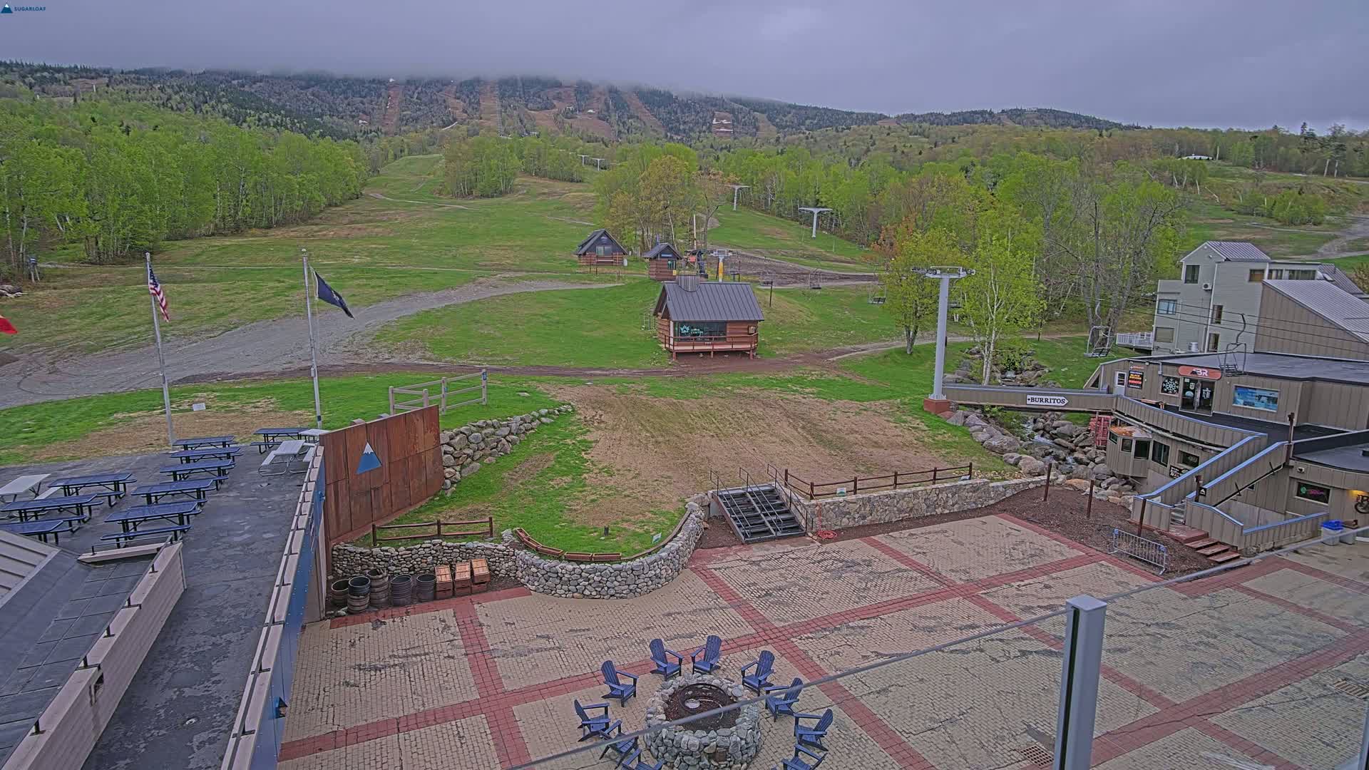 An outdoor paved patio area with picnic tables and a fire pit, overlooking a grassy ski slope with small buildings and a chairlift, under an overcast sky.
