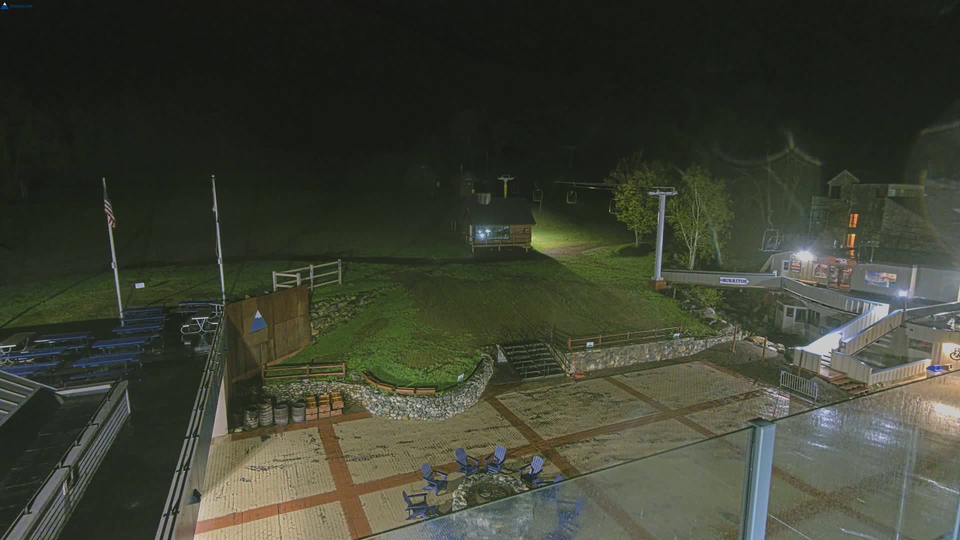 A nighttime view of a ski resort base area with a paved plaza, fire pit, and several buildings under partly cloudy skies.
