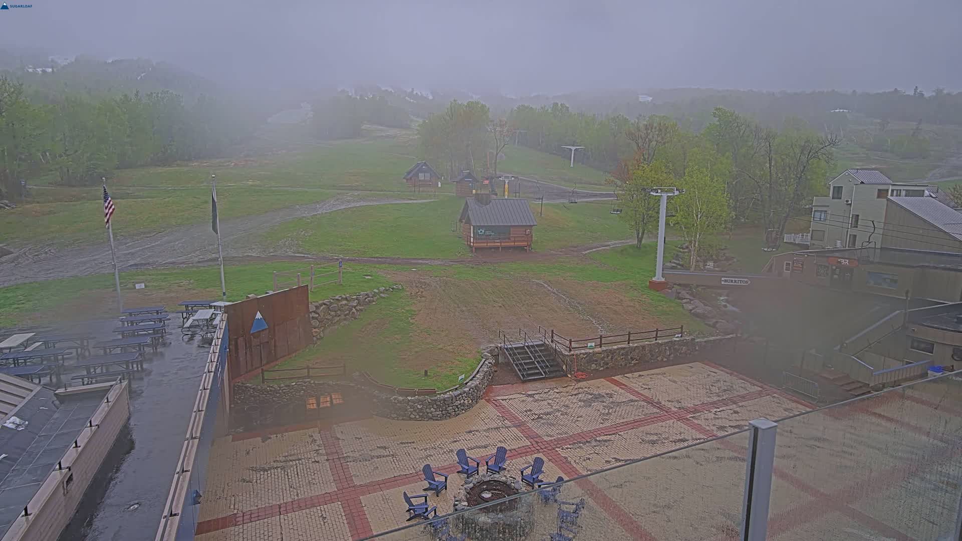 A rainy, foggy view of a ski resort base area showing buildings, picnic tables, a fire pit with chairs, and a ski lift.