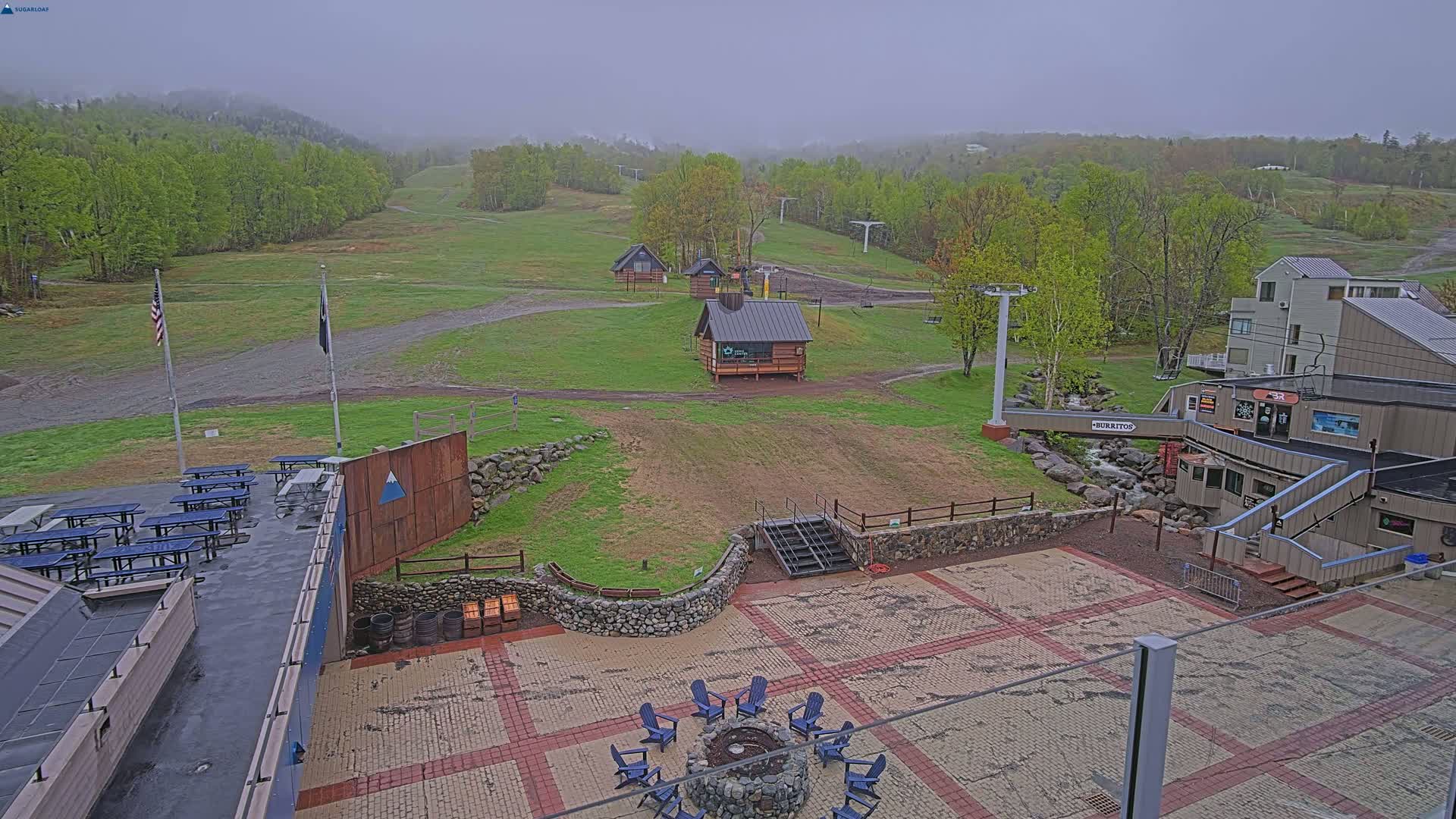 An overcast view of a ski resort's base area, showing a paved patio with a fire pit and seating, picnic tables, small buildings, and a ski lift in the distance.