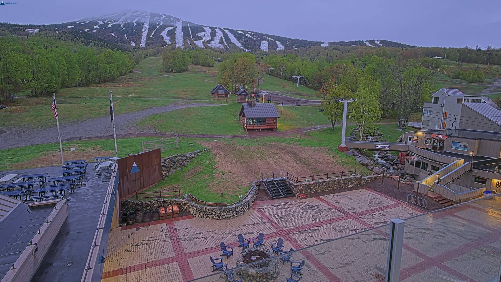 A mountain ski resort base area with a paved plaza, picnic tables, small buildings, and a fire pit, overlooks a mountain with some remaining snow under an overcast sky.