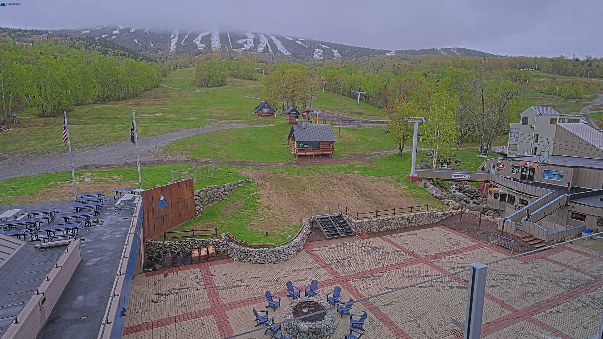 A mostly clear, cool day shows a ski resort base area with picnic tables, small buildings, a fire pit, and a paved patio area; in the background, a mountain with visible ski runs is partly covered in snow.
