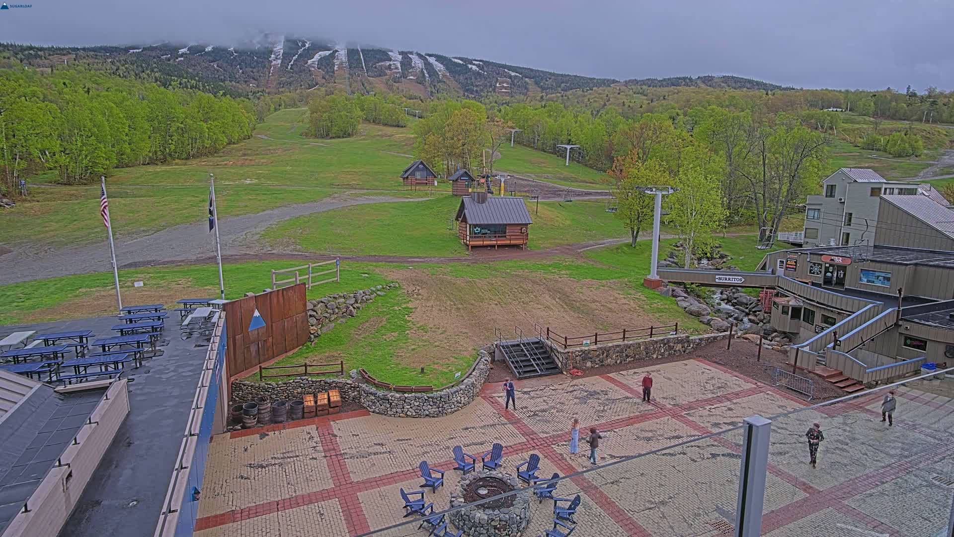 A ski resort base area on an overcast day, showing a paved plaza with picnic tables, a fire pit, small buildings, and a ski lift in the background.