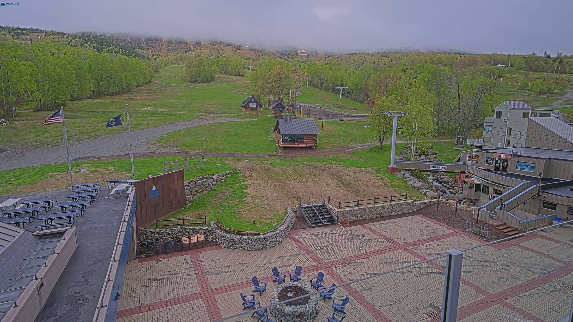 A mostly clear, overcast day reveals a ski resort with picnic tables, a fire pit, small buildings, a chairlift, and a paved patio area, all nestled within a green landscape of trees and hills.