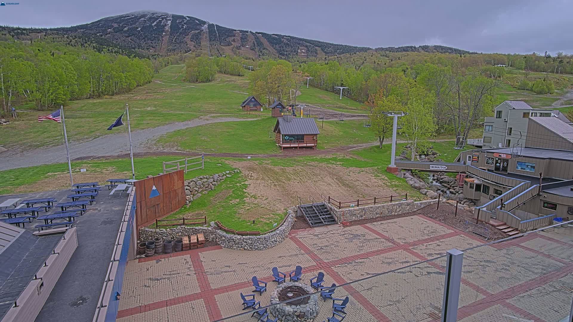 An outdoor patio area with picnic tables, a fire pit, and a small building overlooks a grassy ski slope and mountain under an overcast sky.
