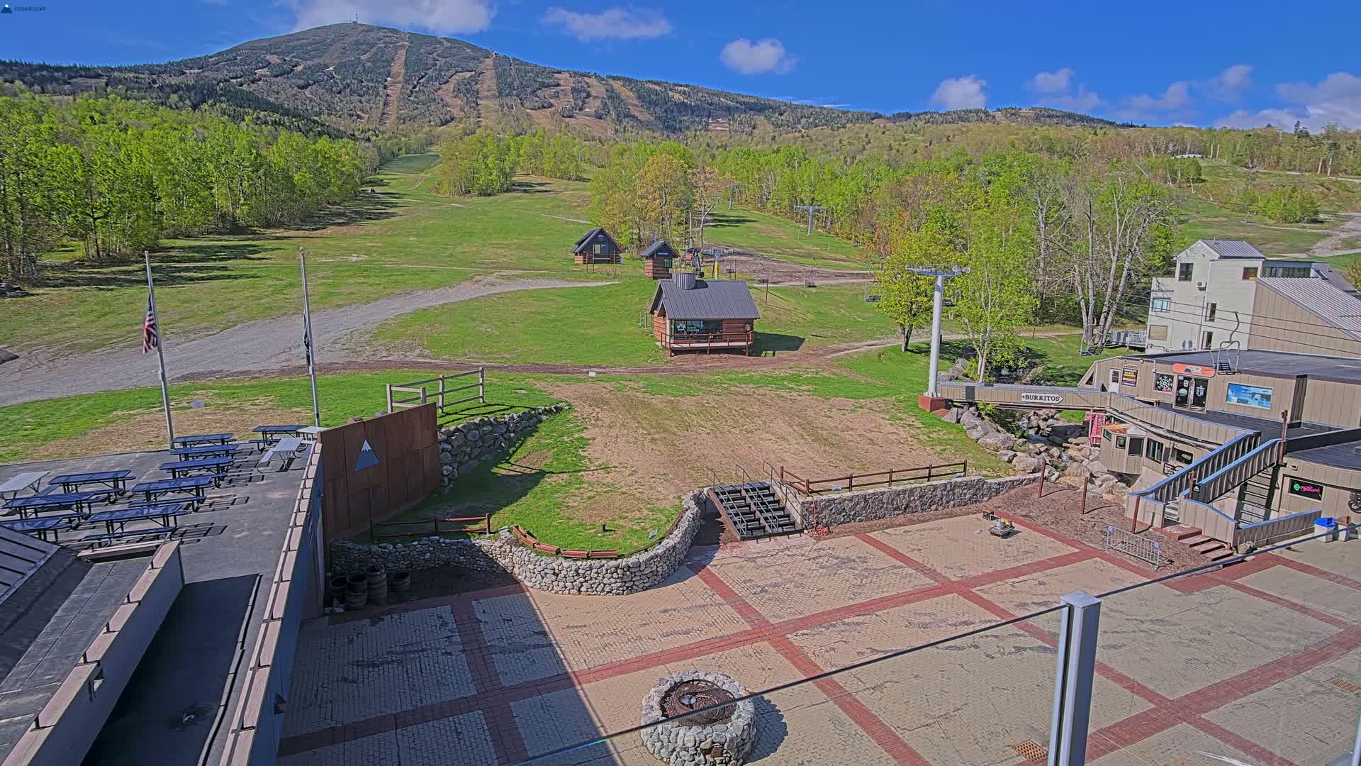 A mountainside ski resort area on a sunny day features a paved plaza with picnic tables, small buildings, and a ski lift, all set against a backdrop of a mountain with green trees.