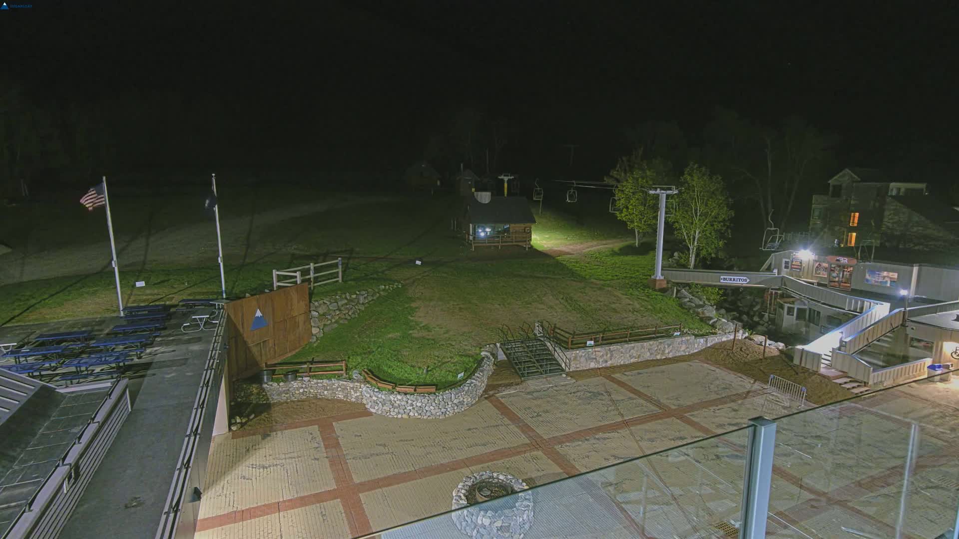 A nighttime view of a ski resort base area with a paved plaza, picnic tables, a small building, a ski lift, and a stone retaining wall, illuminated by artificial lights.