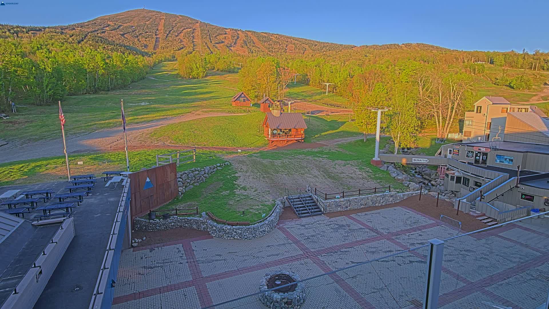 A ski resort base area with several buildings, picnic tables, and a paved plaza under a clear, sunny sky, with a large mountain in the background.