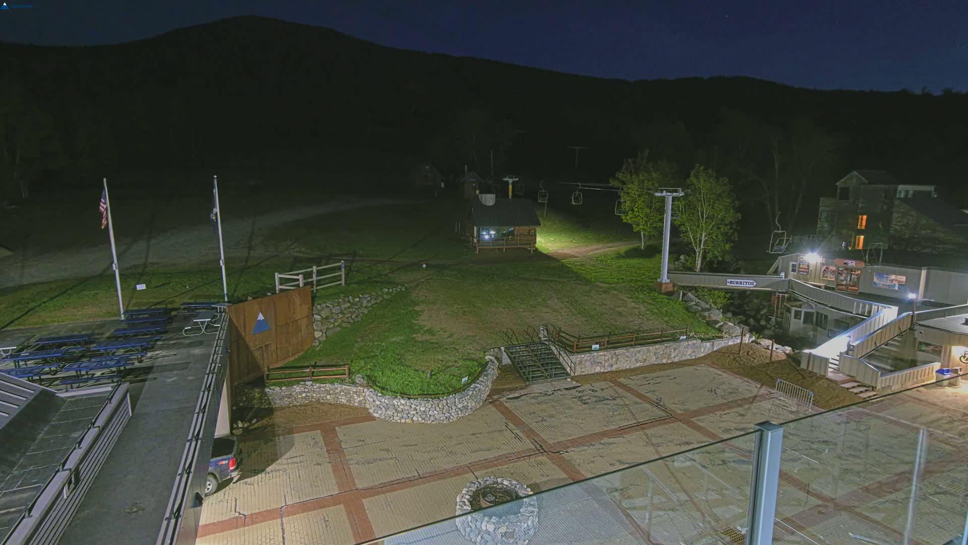 A nighttime view of a ski resort base area, illuminated by artificial lights, showing a paved plaza, a small building, a chairlift, and a grassy area against a dark mountain backdrop.