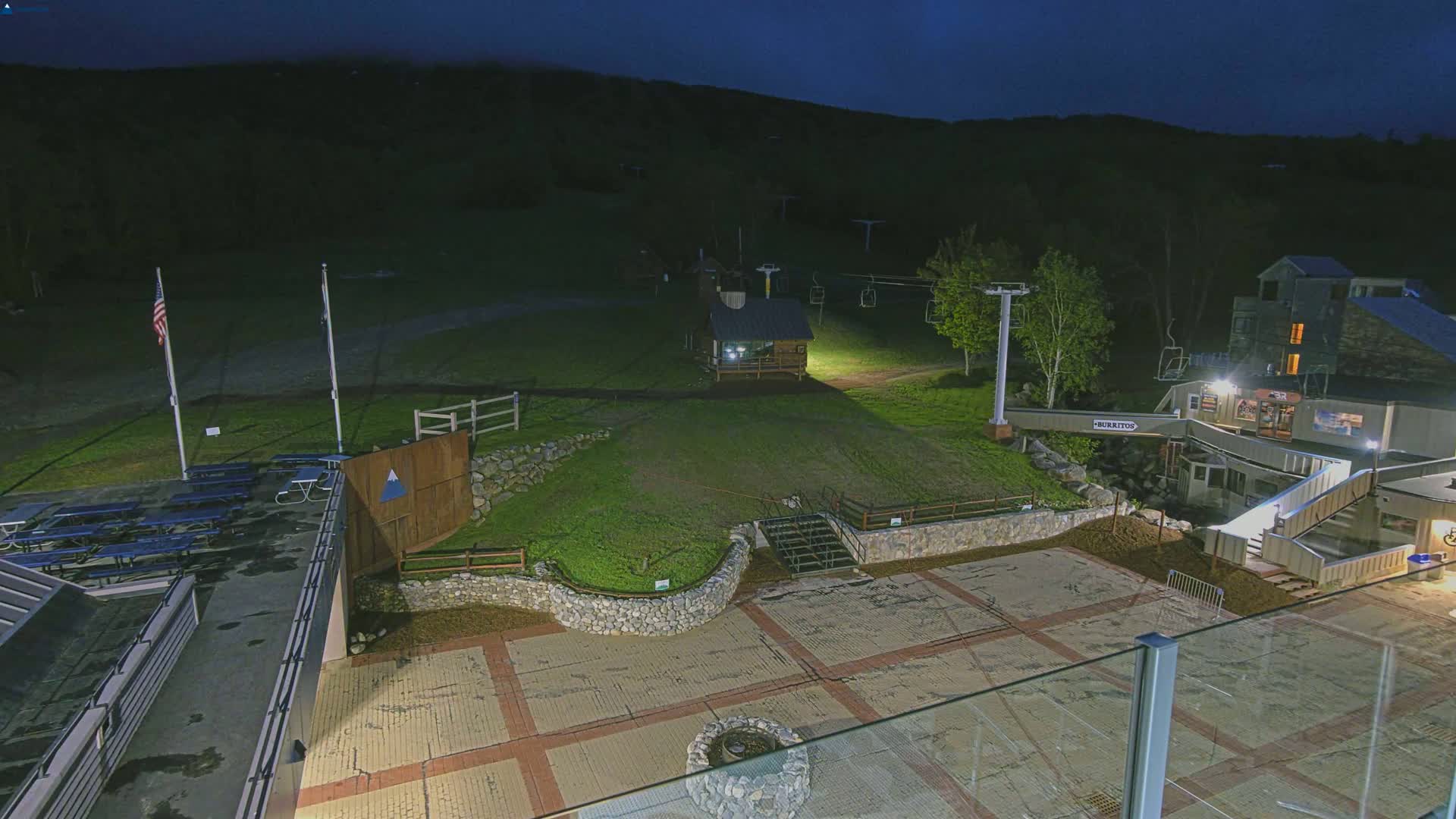 A nighttime view of a ski resort base area with picnic tables, a small building, ski lifts, and a paved plaza illuminated by artificial lights, all set against a dark, hilly background.