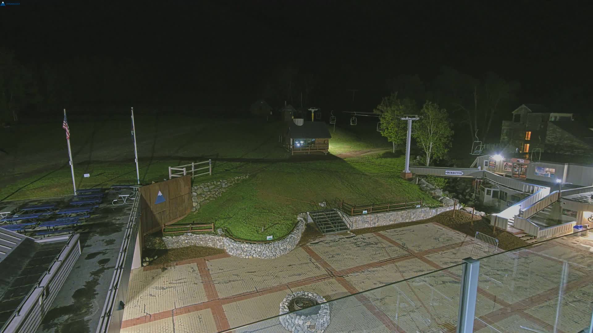 A nighttime view of an outdoor ski resort area with a paved plaza, grassy hills, a ski lift, picnic tables, and a building illuminated by artificial lights.